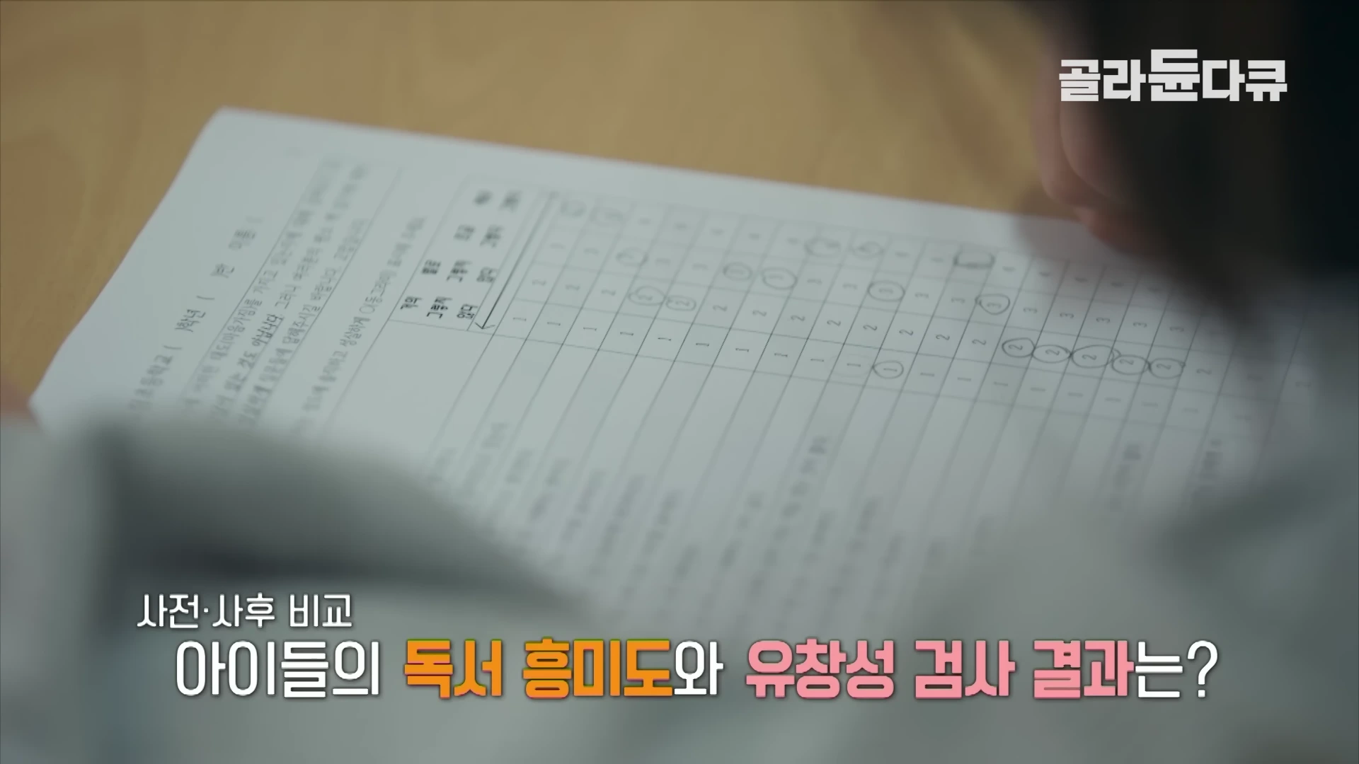 책상 위에 놓인 설문지 종이에 연필로 답변을 체크하고 있는 학생의 모습이 위에서 내려다보는 시점으로 촬영되어 있다. 화면 하단에는 자막으로 '사전·사후 비교 아이들의 독서 흥미도와 유창성 검사 결과는?'이라는 문구가 적혀 있다.