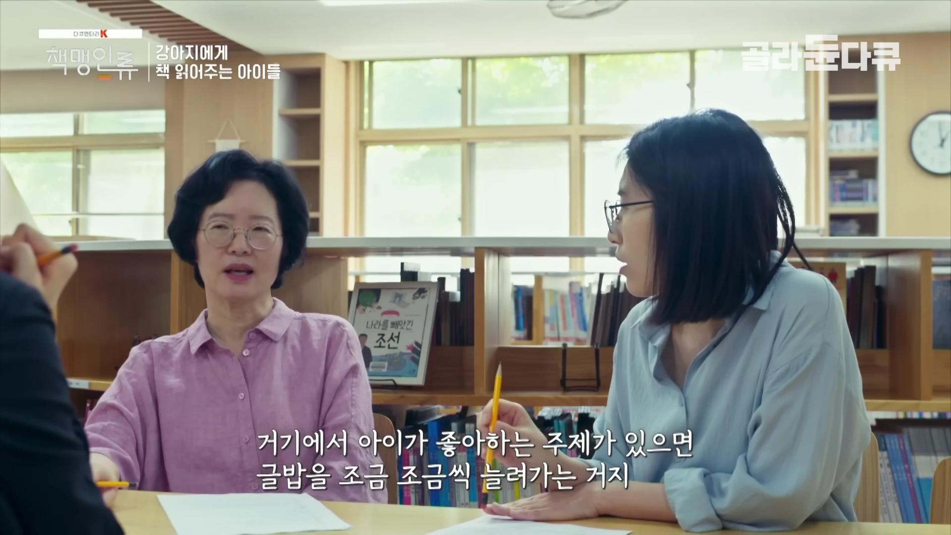 도서관 같은 실내에서 두 여성이 테이블을 사이에 두고 마주 앉아 대화를 나누고 있으며, 자막이 화면 하단에 표시된 영상 캡처 화면입니다.