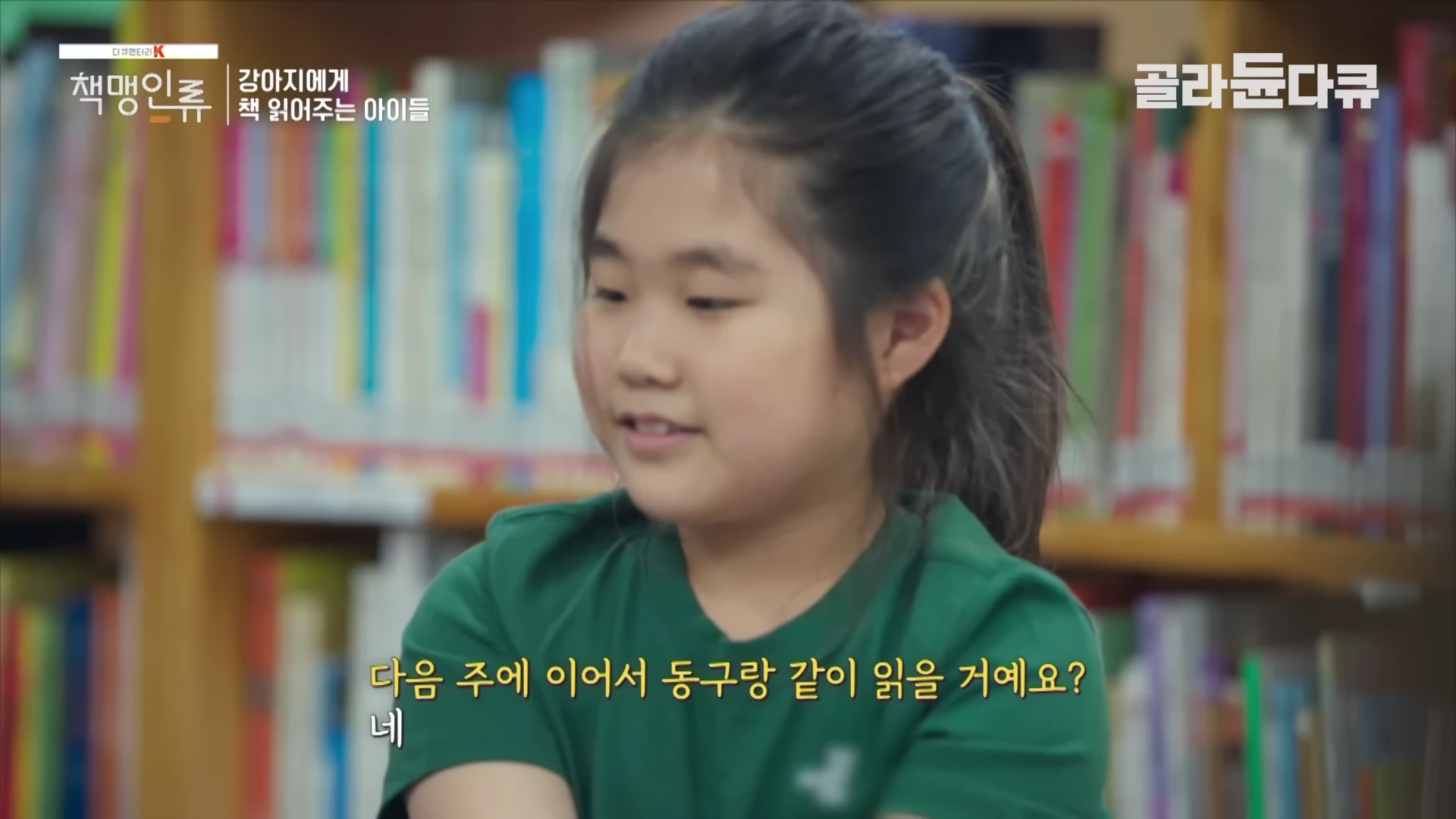 도서관 책장 앞에서 초록색 티셔츠를 입은 여자아이가 강아지에게 책을 읽어주며 대화하고 있는 방송 화면