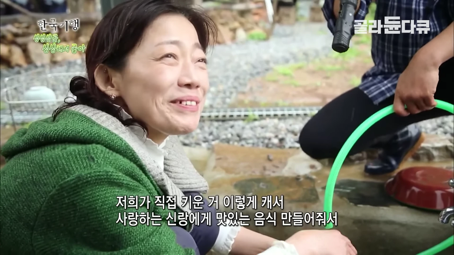 한 여성이 녹색 옷을 입고 밝게 미소 지으며 누군가와 대화하고 있고, 옆에는 물을 뿌리는 호스가 보입니다.