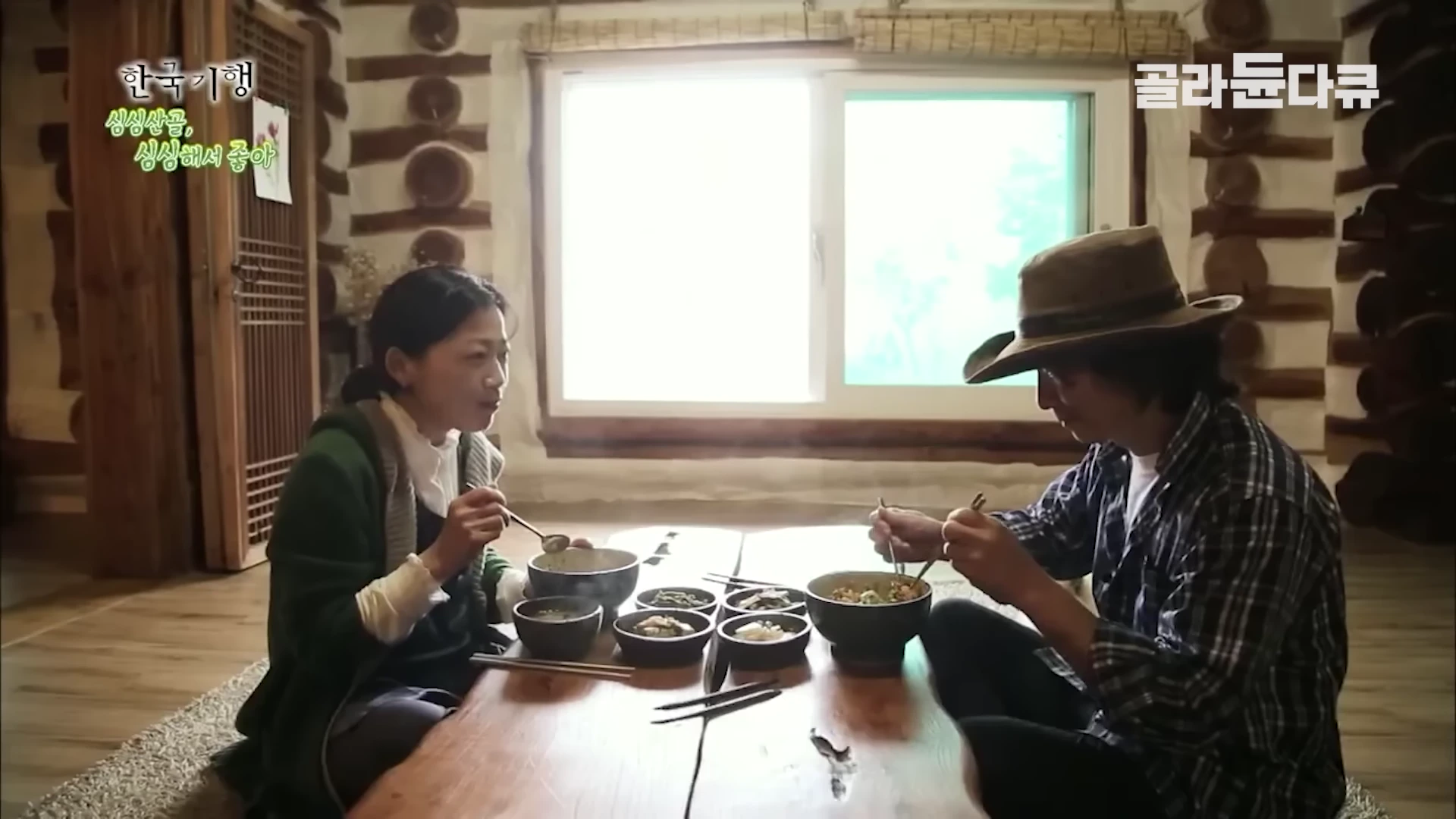 나무 벽으로 된 실내에서 모자를 쓴 남성과 여성이 낮은 나무 식탁에 앉아 식사를 하고 있다.