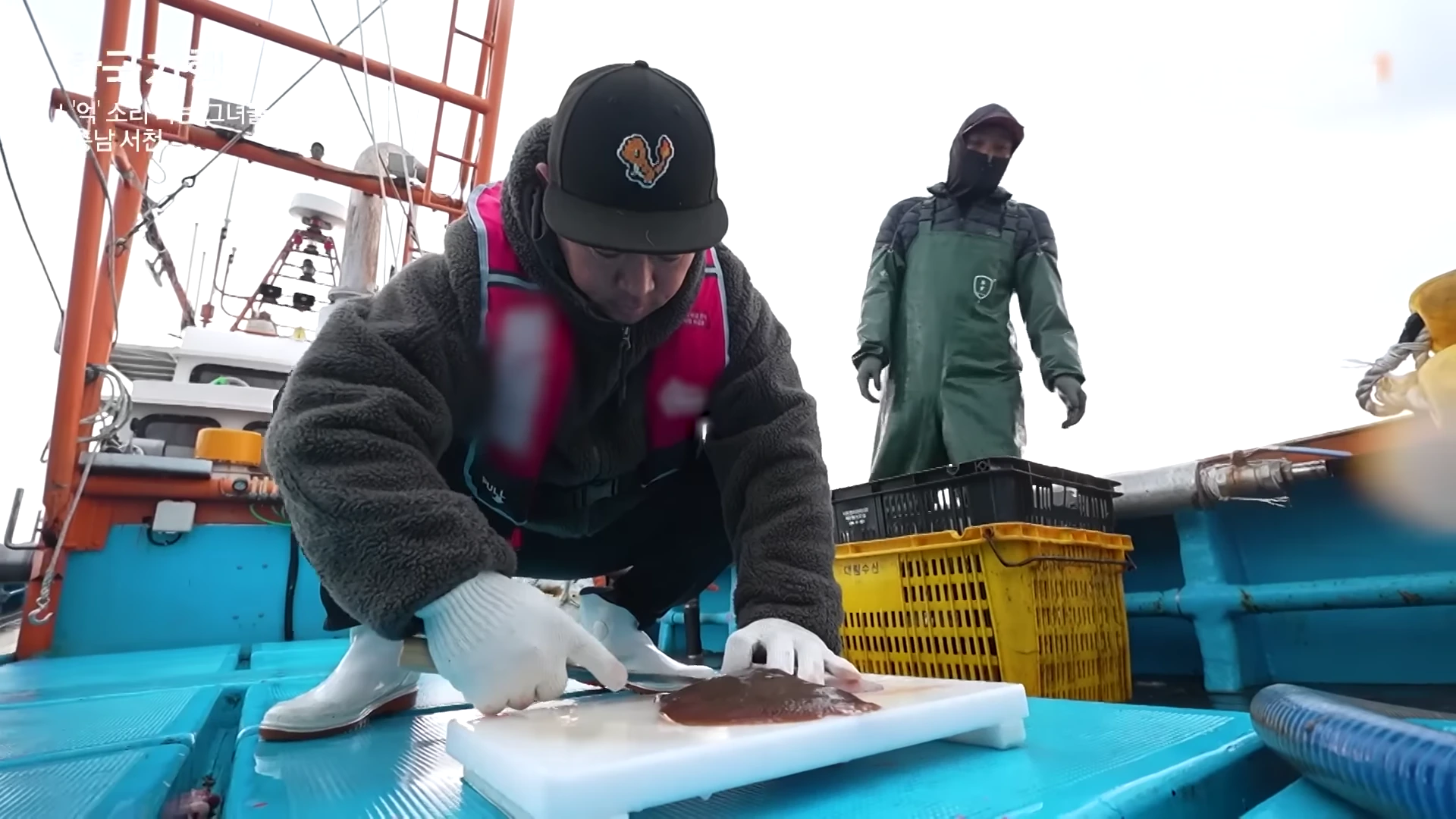 모자와 작업복을 입은 한 남성이 배 위에서 흰 도마 위에 간재미를 올려놓고 칼로 손질하고 있으며 뒤쪽에는 다른 작업자가 서 있다.