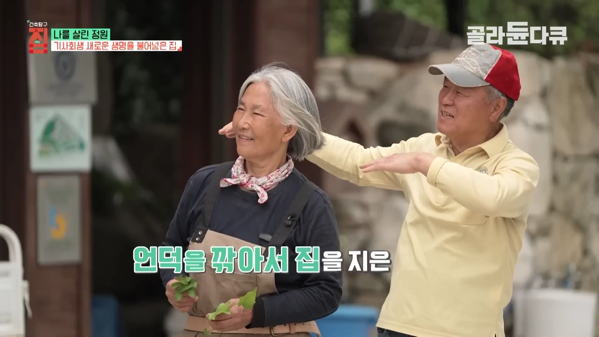 순천 흙집 정원에서 중년 부부가 함께 서서 대화를 나누고 있으며, 여성은 앞치마를 두르고 식물을 들고 있고 남성은 손짓하며 설명하는 모습.