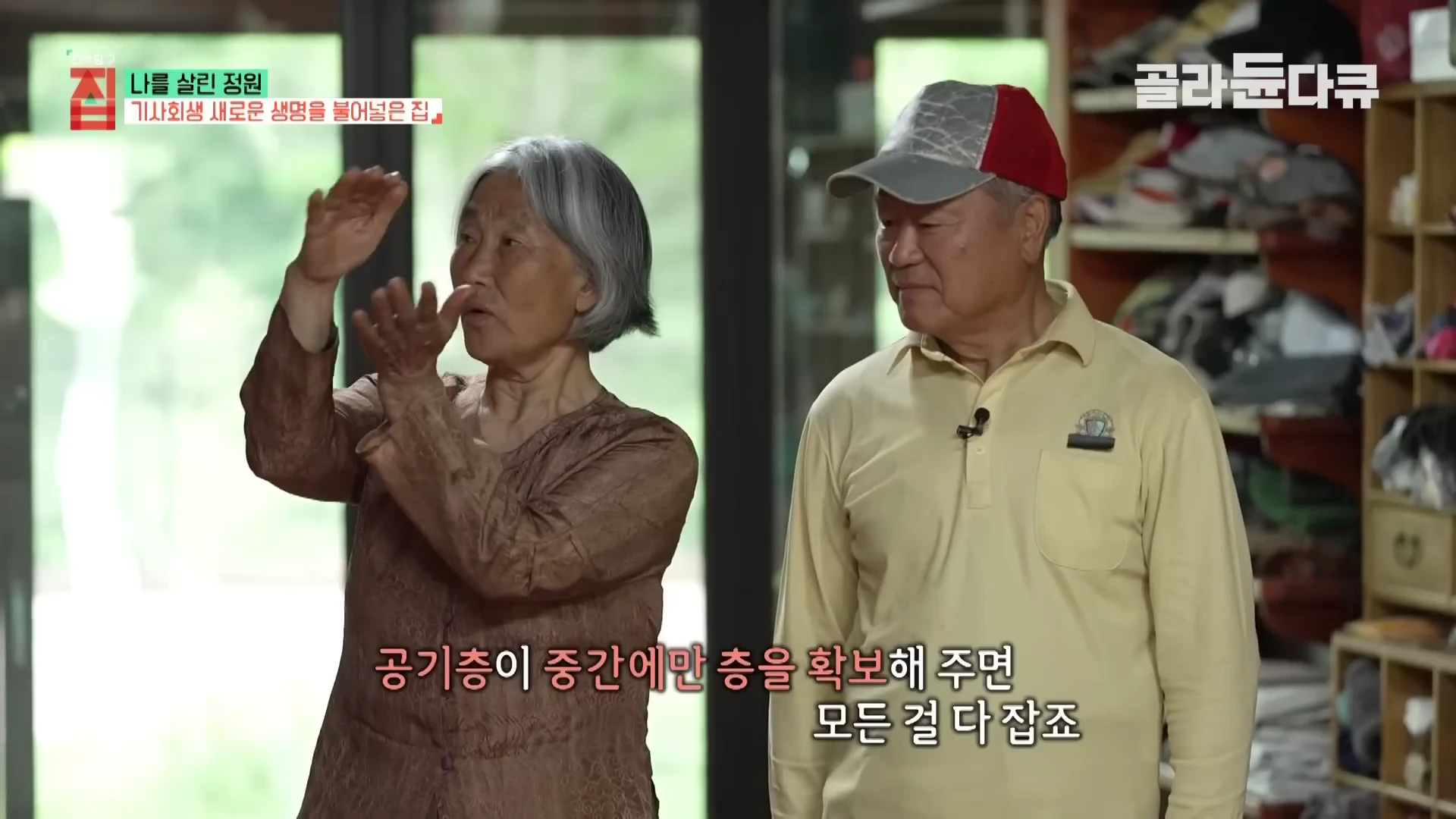 실내에서 중년 부부가 서서 대화를 나누고 있으며, 아내가 손을 움직이며 집 구조에 대해 설명하고 있다.