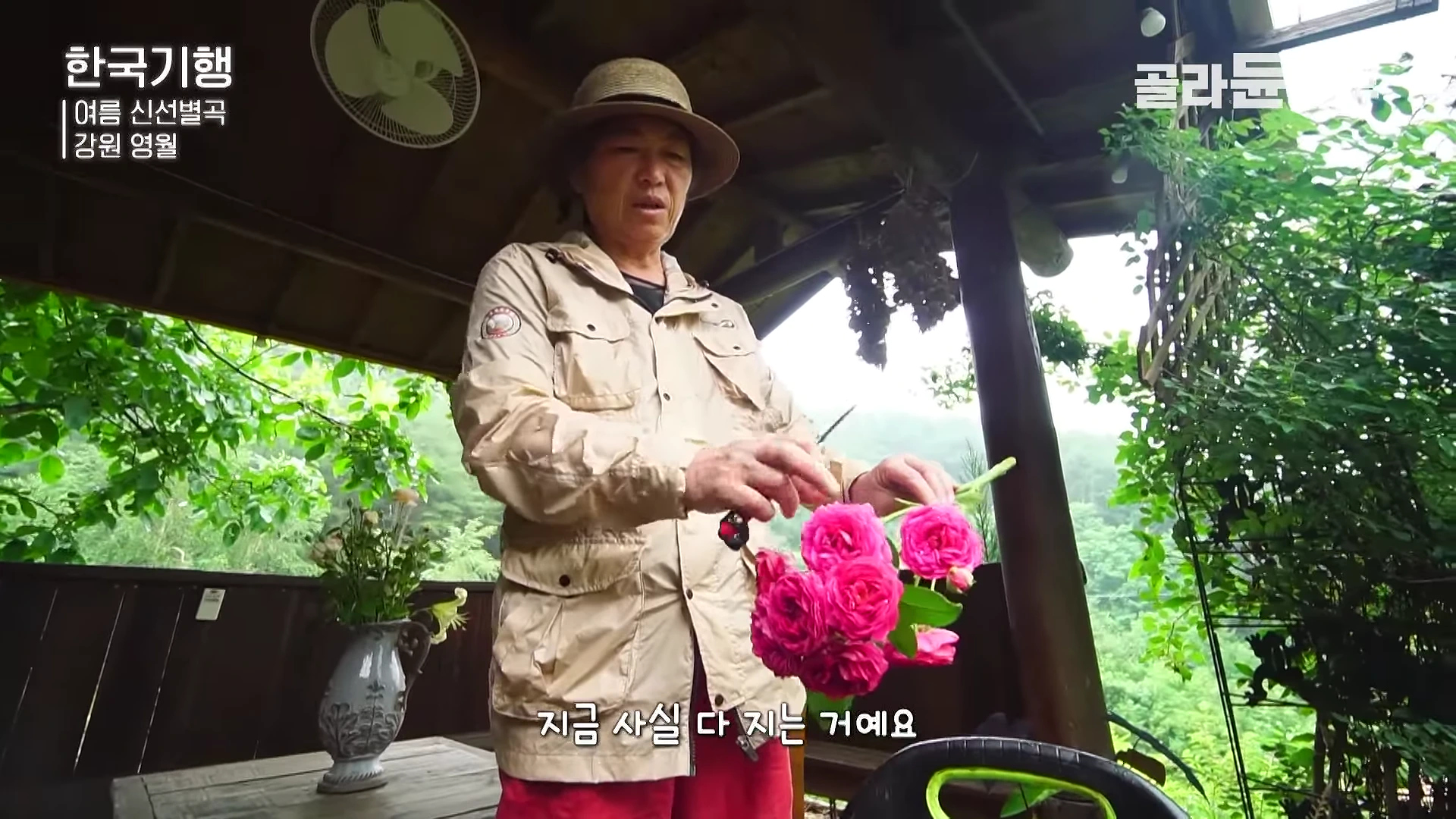 챙이 있는 모자를 쓰고 아웃도어 재킷을 입은 중년 여성이 야외 정자에서 분홍색 꽃송이들을 다듬으며 이야기하고 있다.
