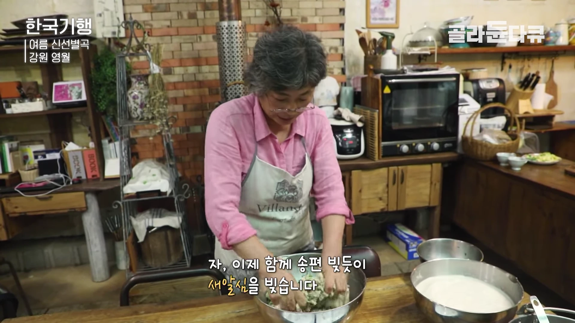 부엌에서 분홍색 셔츠와 앞치마를 입은 여성이 스테인리스 볼에 담긴 감자 반죽을 손으로 빚고 있다.