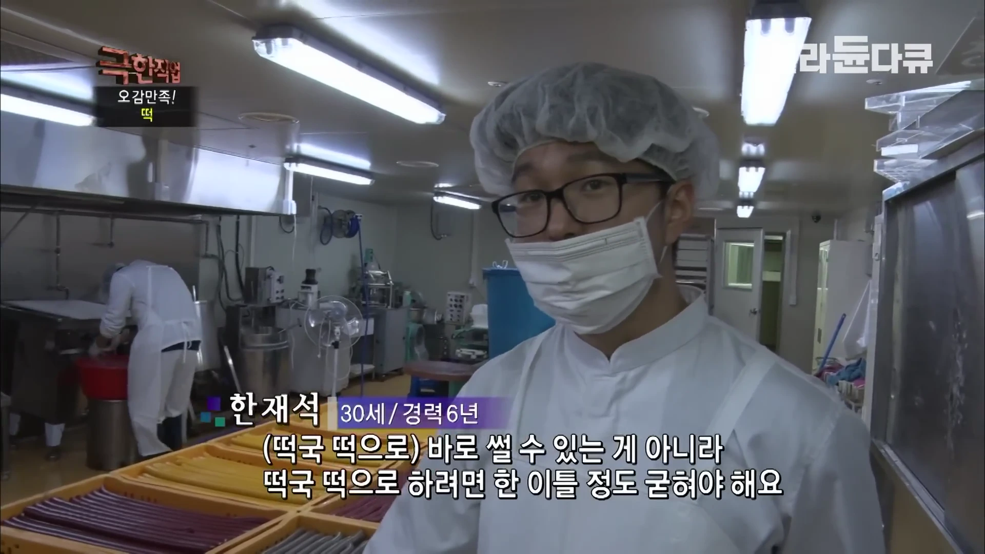 위생모와 마스크를 착용한 젊은 작업자가 작업장에서 카메라를 바라보며 떡 제조 과정을 설명하고 있고, 배경에는 오색 가래떡이 담긴 바구니와 작업 중인 다른 사람이 보입니다.