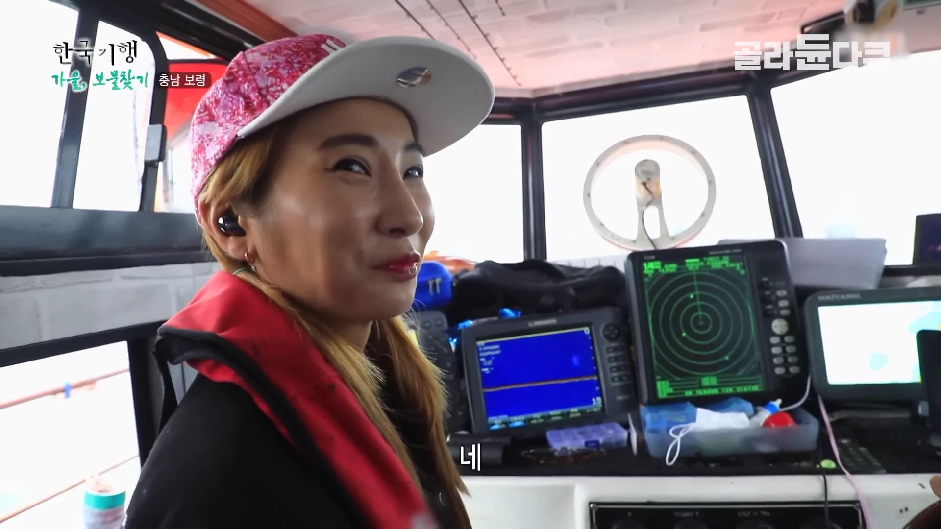배 조타실에서 핑크색 야구 모자를 쓴 여성이 밝은 표정으로 카메라를 바라보고 있고 주변에는 항해용 레이더와 모니터가 설치되어 있습니다.