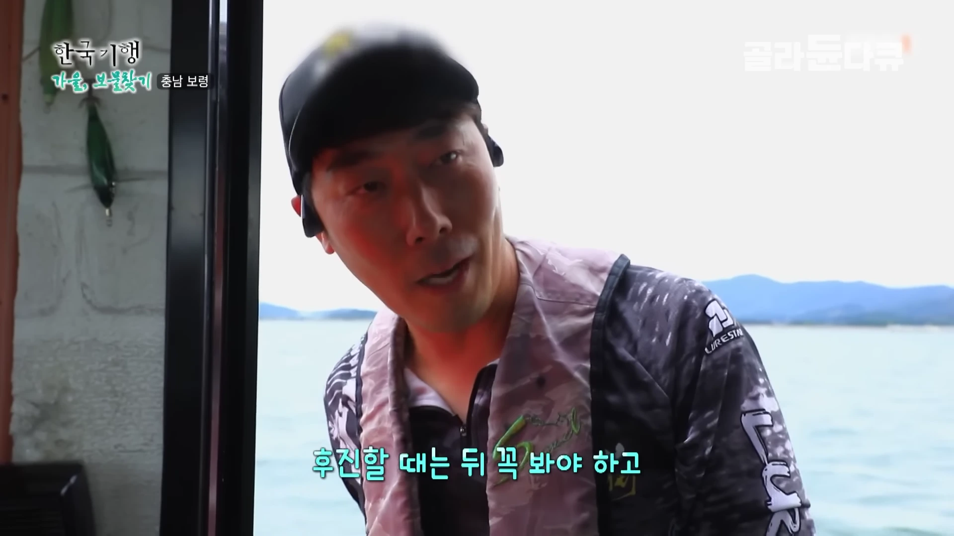 낚싯배 조종석에서 구명조끼를 입은 남성이 정면을 바라보며 조언을 건네고 있는 모습