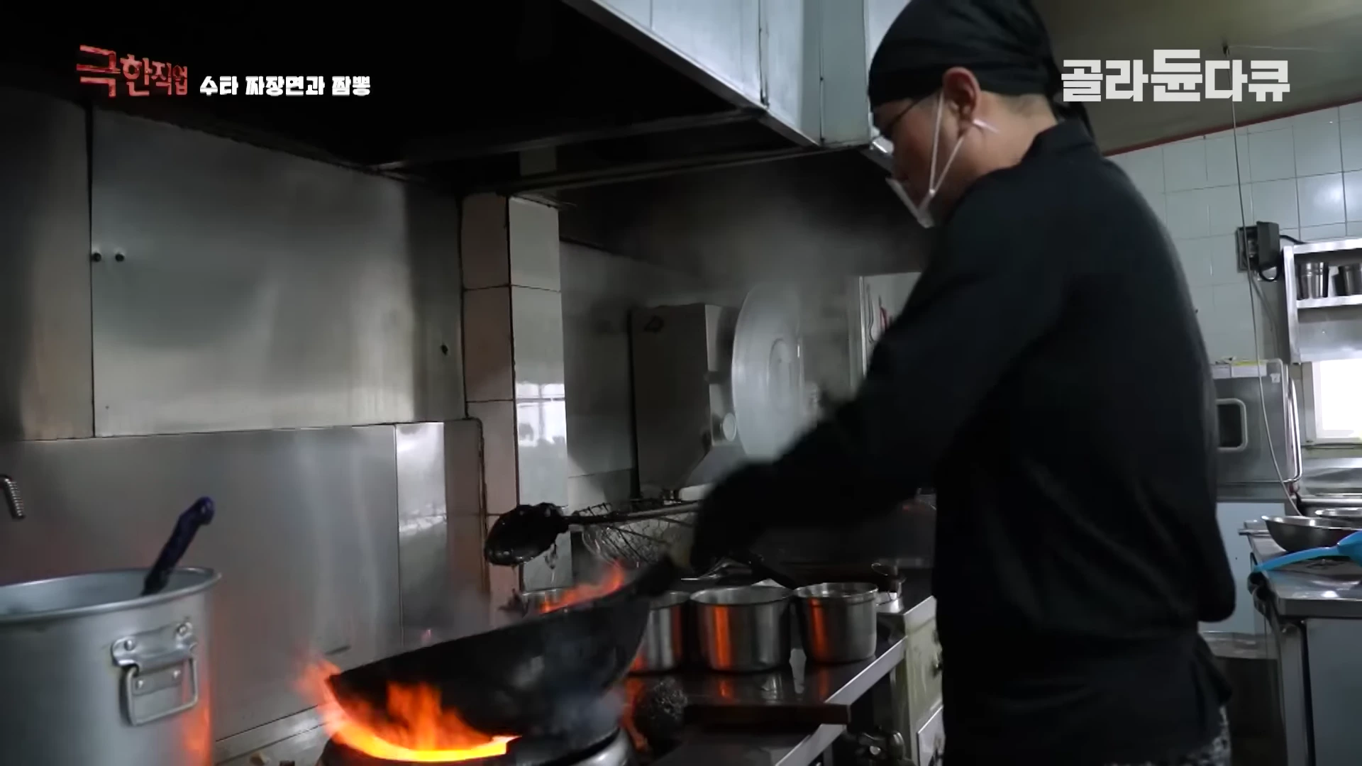 검은색 작업복을 입고 마스크를 쓴 주방장이 뜨거운 불길이 치솟는 웍을 잡고 요리하는 모습