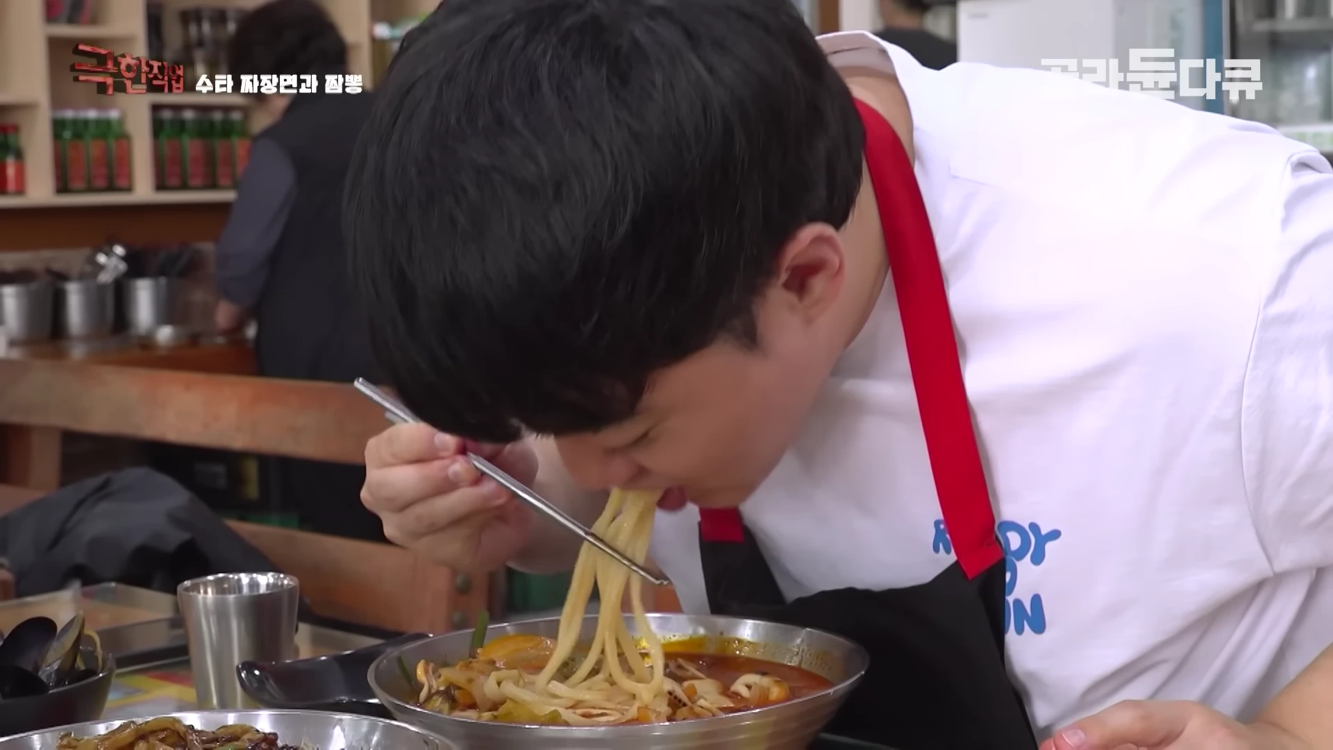 주방에서 흰색 상의와 검은색 앞치마를 입은 요리사가 젓가락으로 갓 만든 짬뽕 면을 들어 올려 먹고 있는 모습
