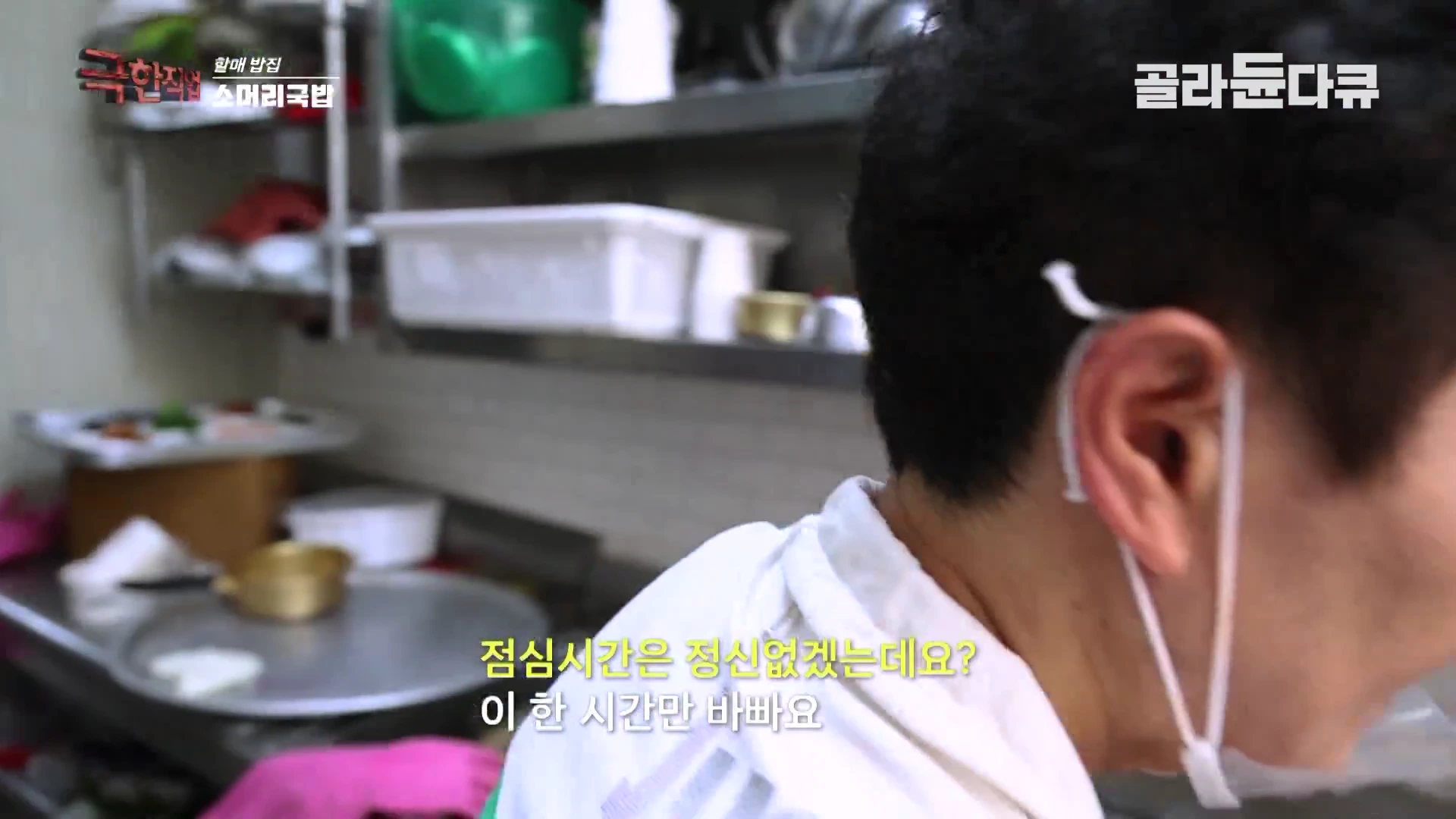 흰색 위생복과 마스크를 착용한 사람이 주방에서 음식을 준비하고 있는 옆모습이며 화면 하단에는 자막이 포함되어 있습니다.