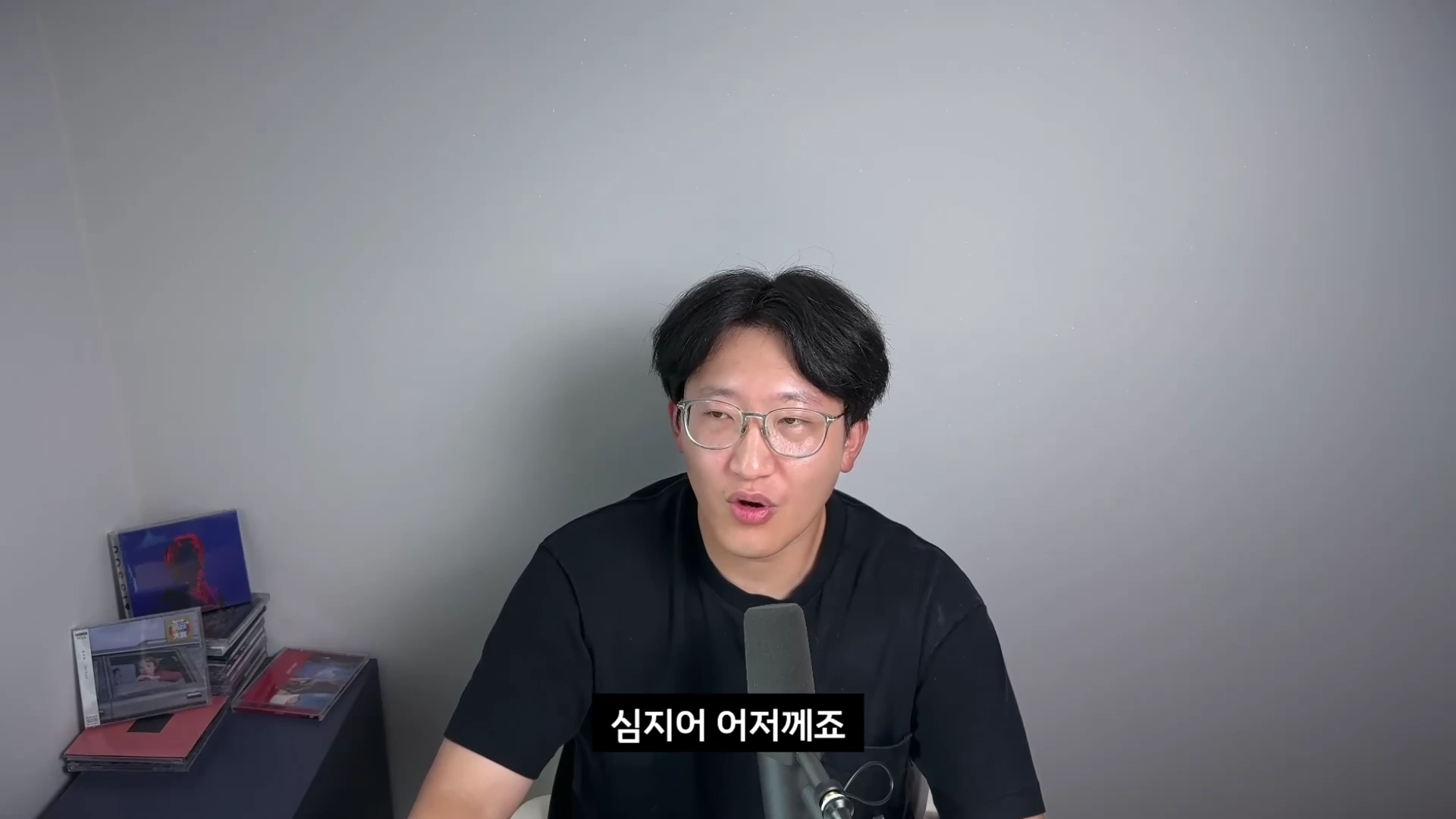 검은색 티셔츠를 입고 안경을 쓴 남성이 마이크 앞에 앉아 정면을 보며 이야기를 하고 있다.