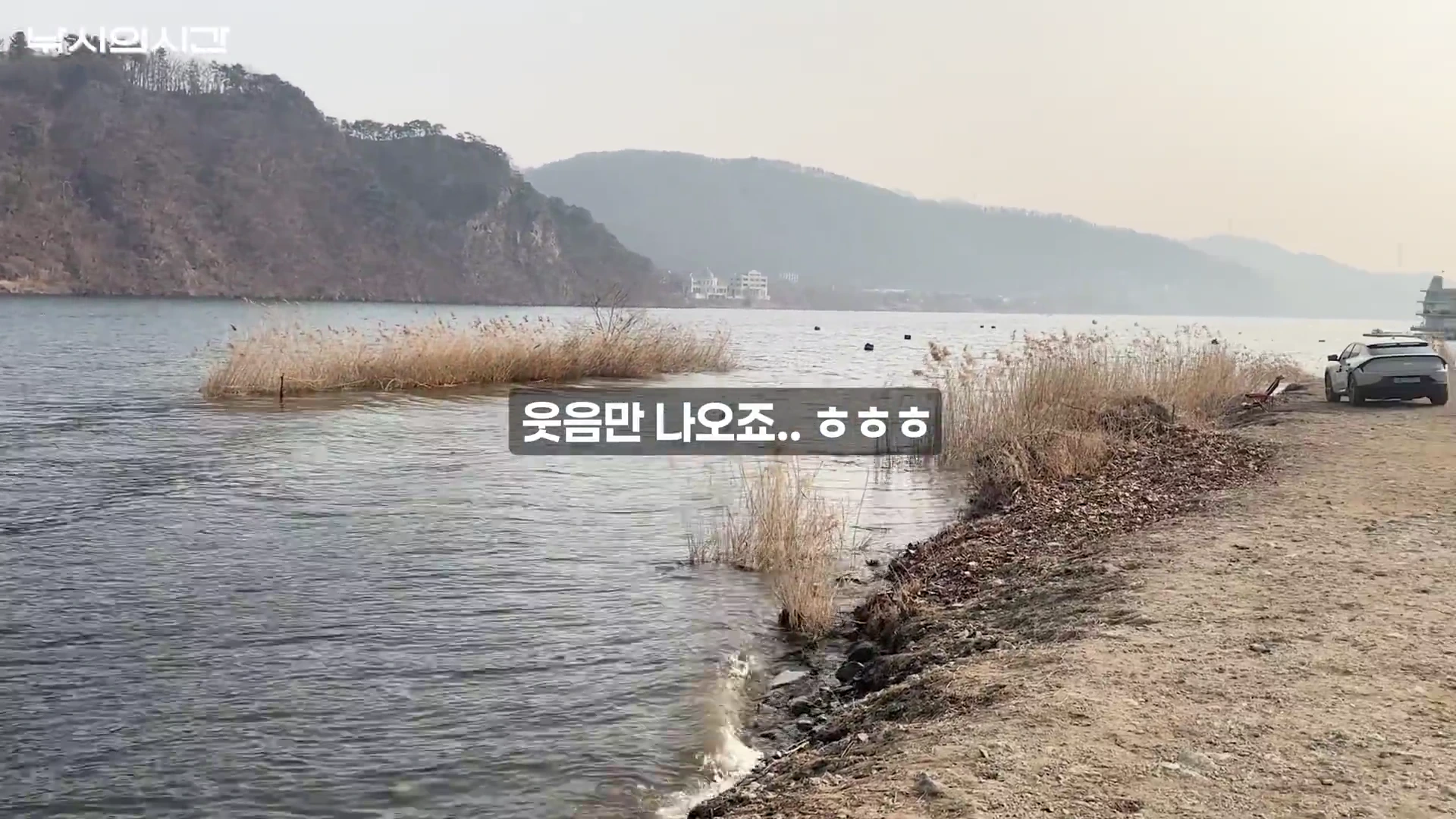 강가에 갈대밭이 펼쳐진 노지 낚시터 풍경으로, 비포장 도로 위에 주차된 차량과 멀리 산이 보이는 평화로운 전경입니다.