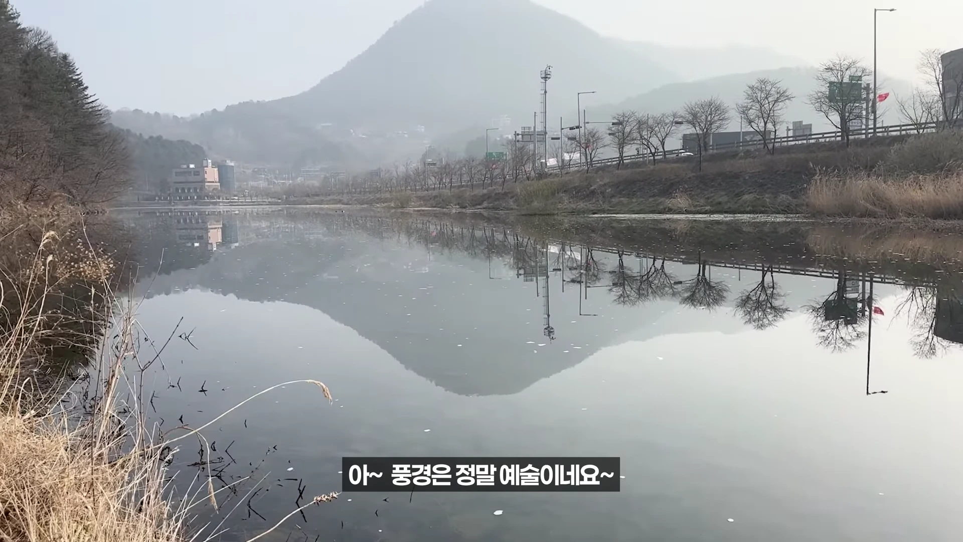 잔잔한 강물에 건너편 산과 가로등이 비치는 하천 풍경이 담긴 영상 캡처 화면이며, 하단에는 한국어 자막이 표시되어 있습니다.