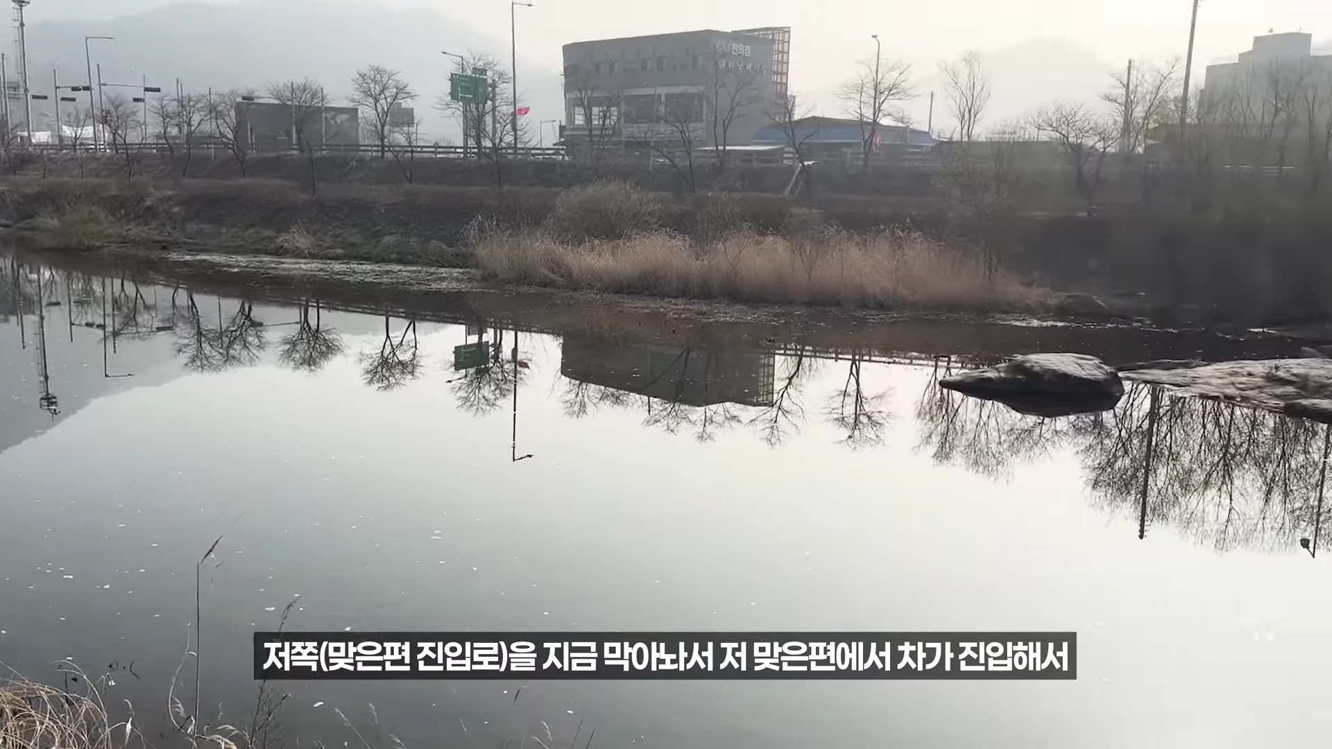 잔잔한 물이 흐르는 강가 풍경으로, 건너편에 건물들과 갈대밭이 보이며 하단에는 자막이 포함된 화면입니다.