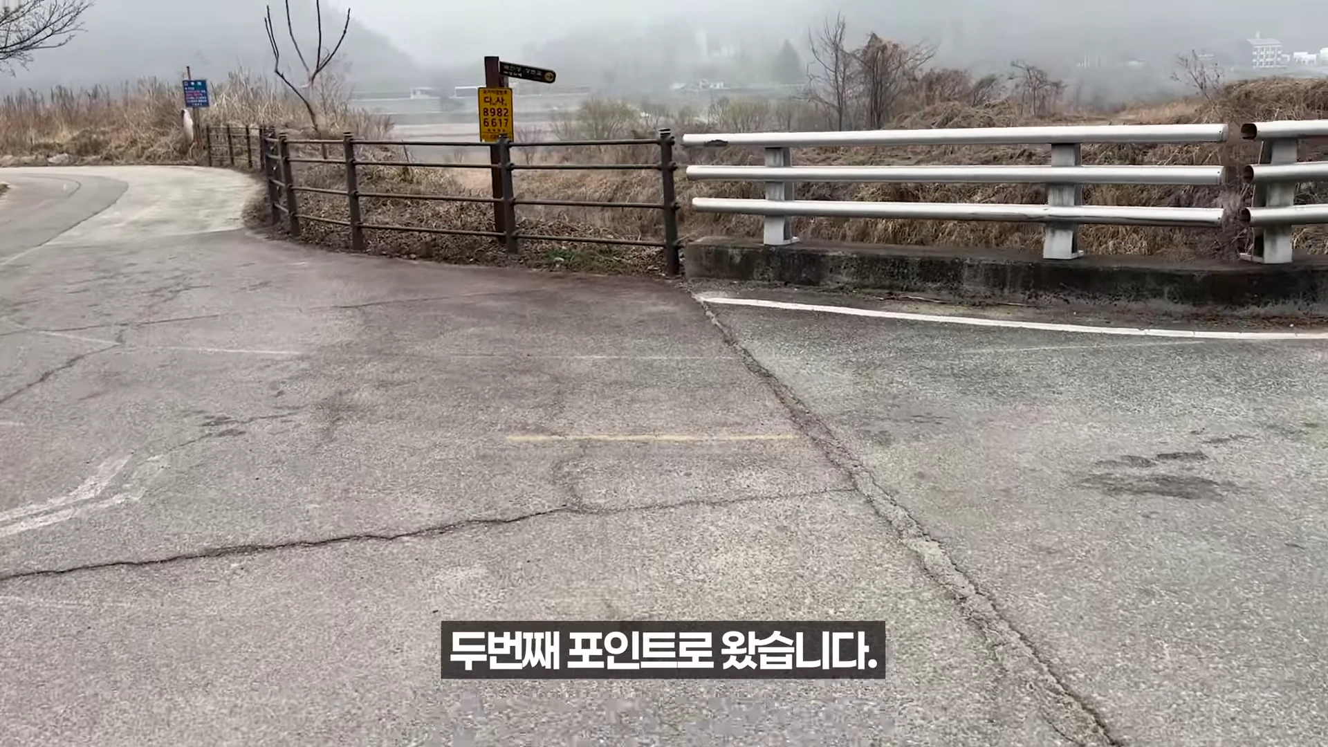 포장된 도로 옆에 설치된 철제 난간과 노란색 표지판이 보이고 그 뒤로 갈색 풀이 우거진 하천변 풍경이 펼쳐진 실외 모습입니다.