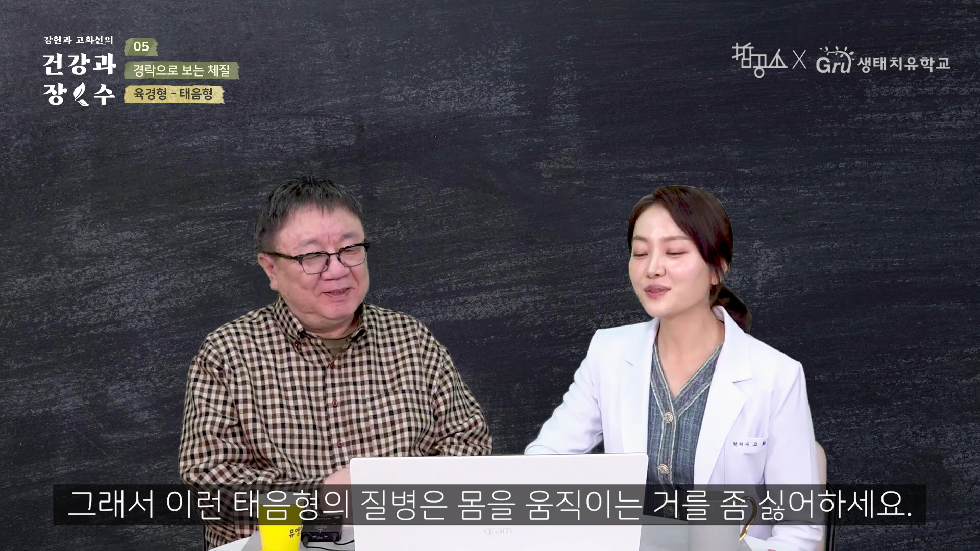 두 사람이 검은색 배경의 스튜디오에서 책상을 사이에 두고 대화를 나누고 있는 모습으로, 왼쪽에는 안경을 쓴 중년 남성이, 오른쪽에는 의사 가운을 입은 여성이 앉아 있다.