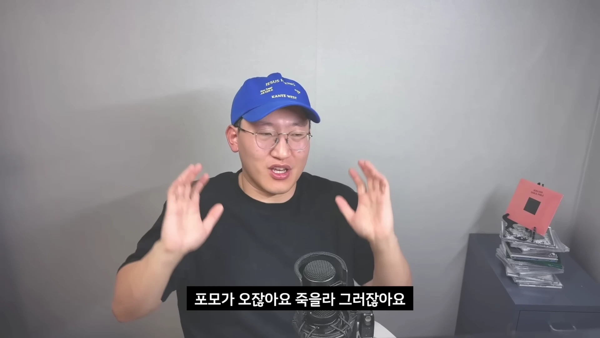 파란색 야구 모자를 쓰고 안경을 쓴 남성이 마이크 앞에서 손을 들어 올리며 이야기를 하고 있고, 화면 하단에는 자막이 있다.