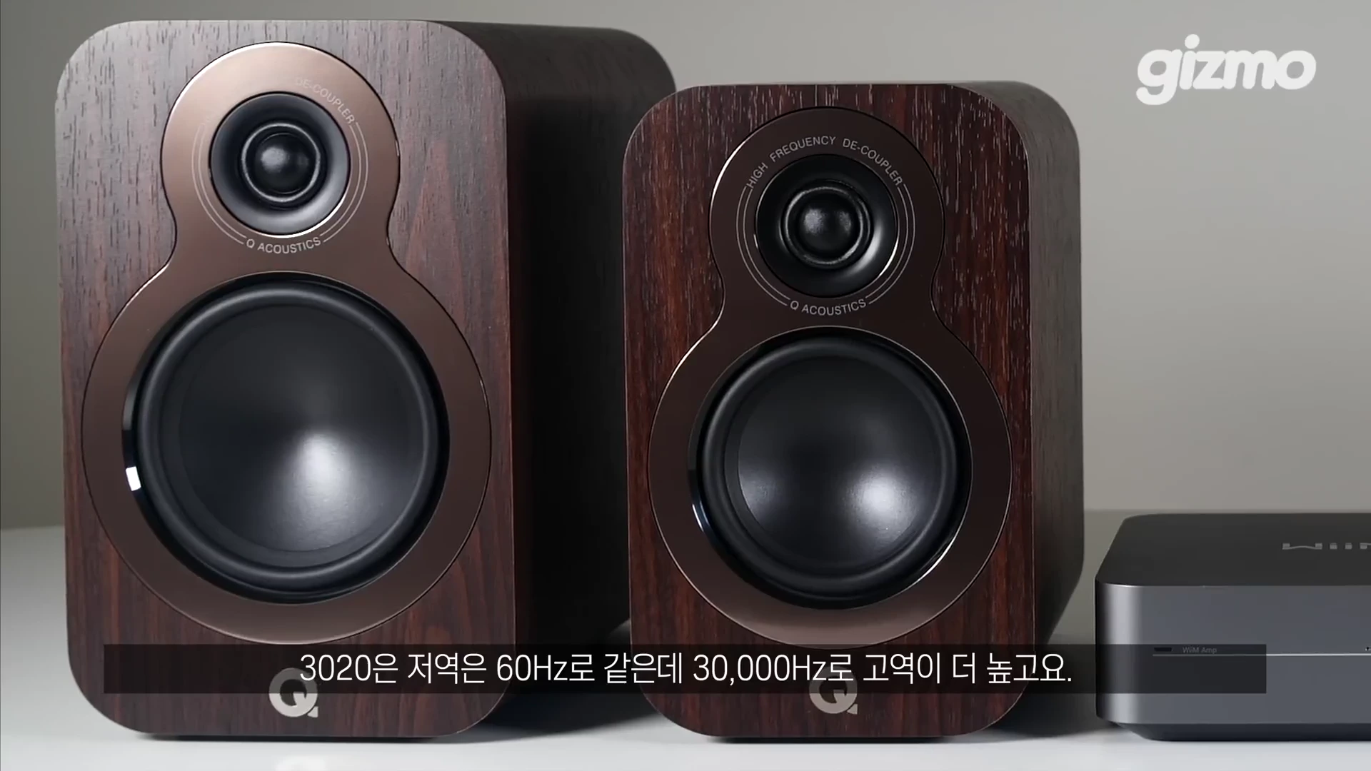 나무 무늬의 북쉘프 스피커 두 대와 그 옆에 놓인 작은 윔앰프 기기가 테이블 위에 배치된 모습.