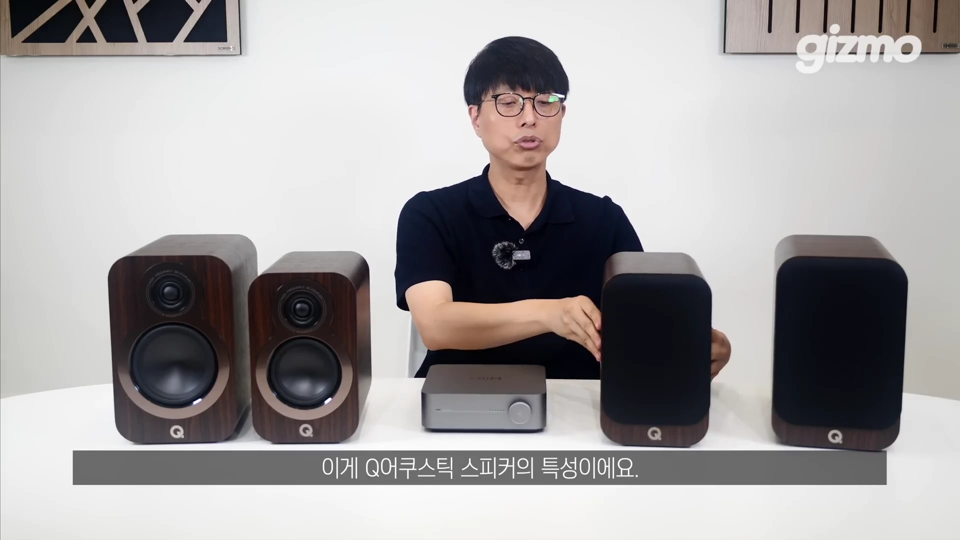 흰 배경의 스튜디오에서 안경을 쓴 남성이 북쉘프 스피커 두 대와 작은 앰프를 앞에 두고 제품을 설명하고 있다.