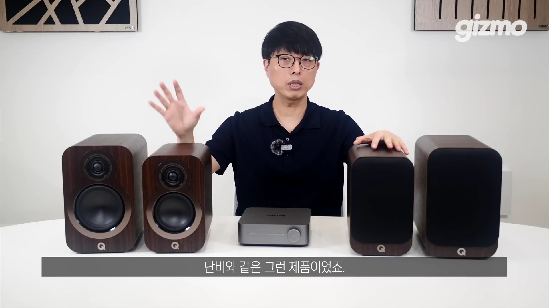 안경을 쓴 남성이 테이블 위에 놓인 윔앰프와 Q어쿠스틱 북쉘프 스피커 세트를 소개하고 있다.