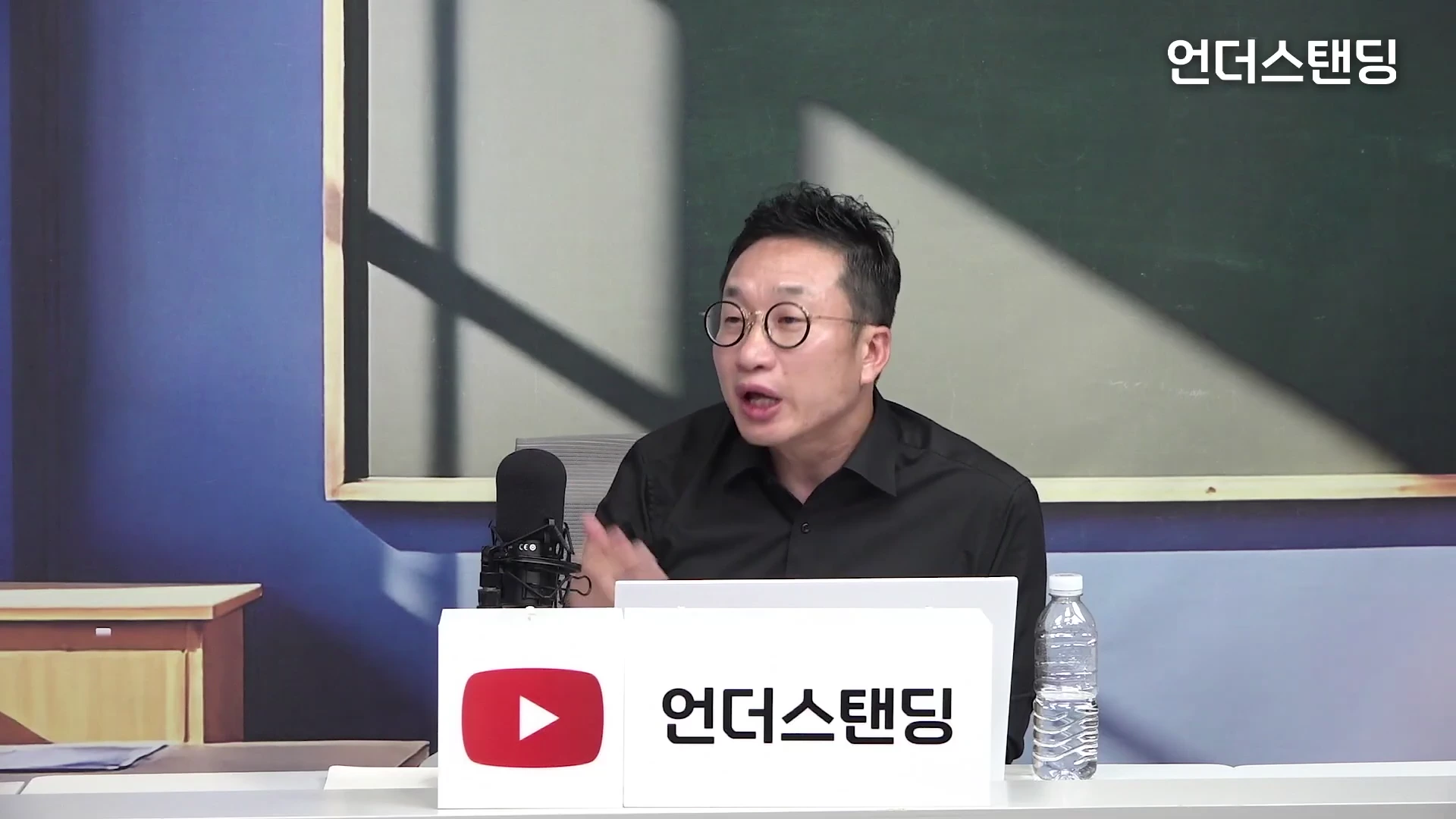 안경을 쓴 남성이 스튜디오 테이블 앞에 앉아 마이크를 앞에 두고 이야기를 나누고 있는 모습
