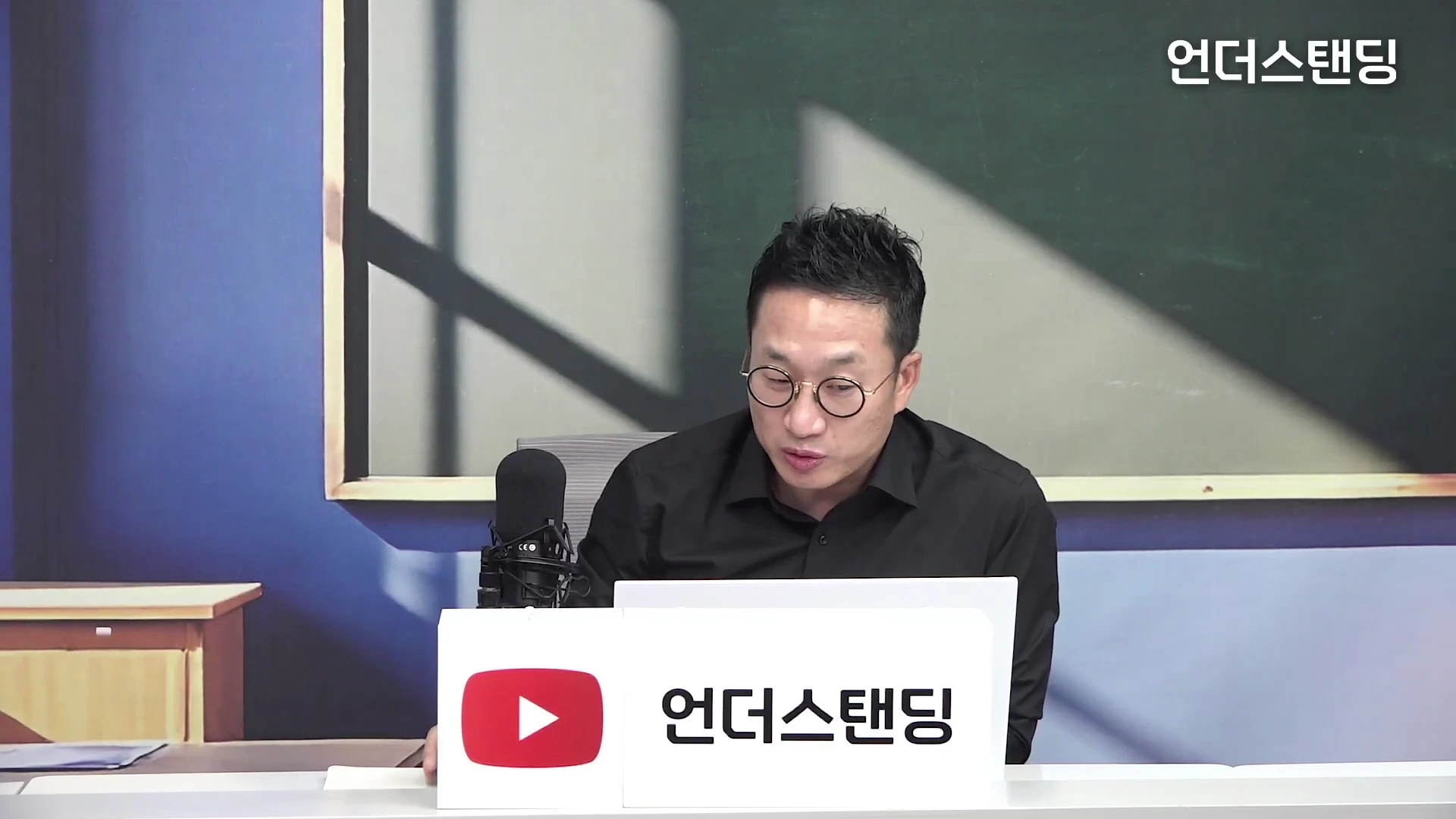 안경을 쓴 남성이 스튜디오 책상 앞에 앉아 마이크를 앞에 두고 이야기를 나누고 있는 모습