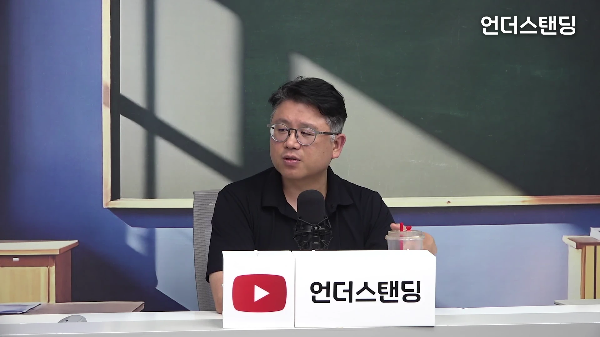 안경을 쓴 중년 남성이 검은색 옷을 입고 마이크 앞에서 이야기를 나누고 있는 스튜디오 인터뷰 장면입니다.
