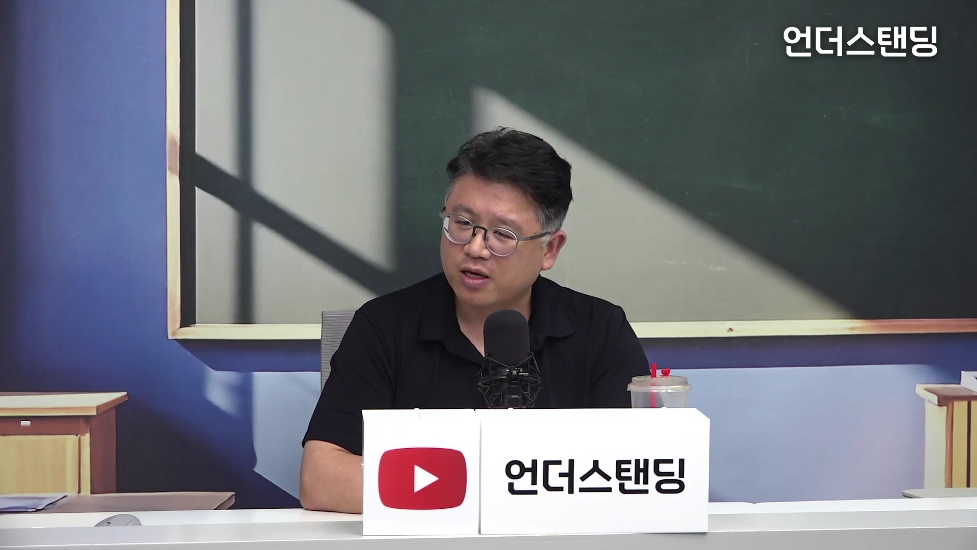 안경을 쓴 중년 남성이 스튜디오 책상에 앉아 마이크 앞에 대고 이야기하는 모습.
