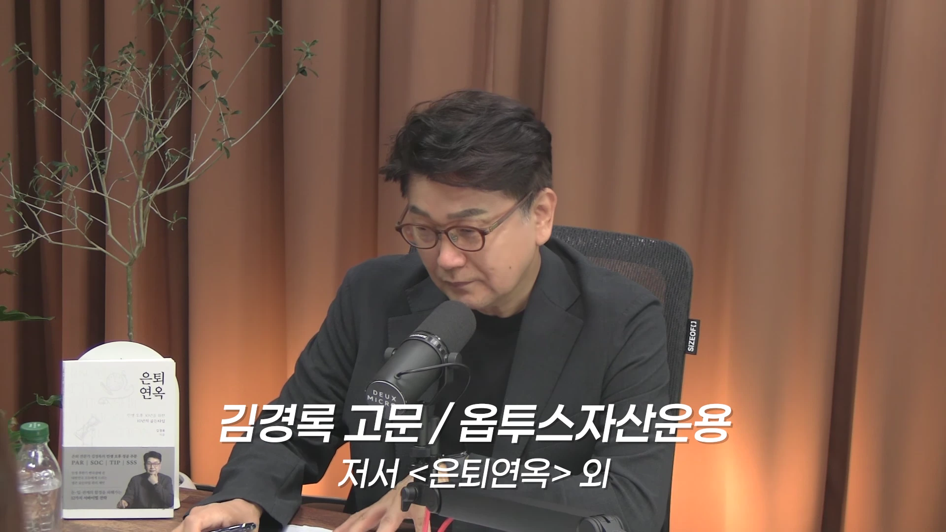 안경을 쓴 중년 남성이 스튜디오에서 마이크 앞에 앉아 인터뷰를 진행하고 있으며, 왼쪽에는 그가 저술한 은퇴 관련 도서가 놓여 있습니다.