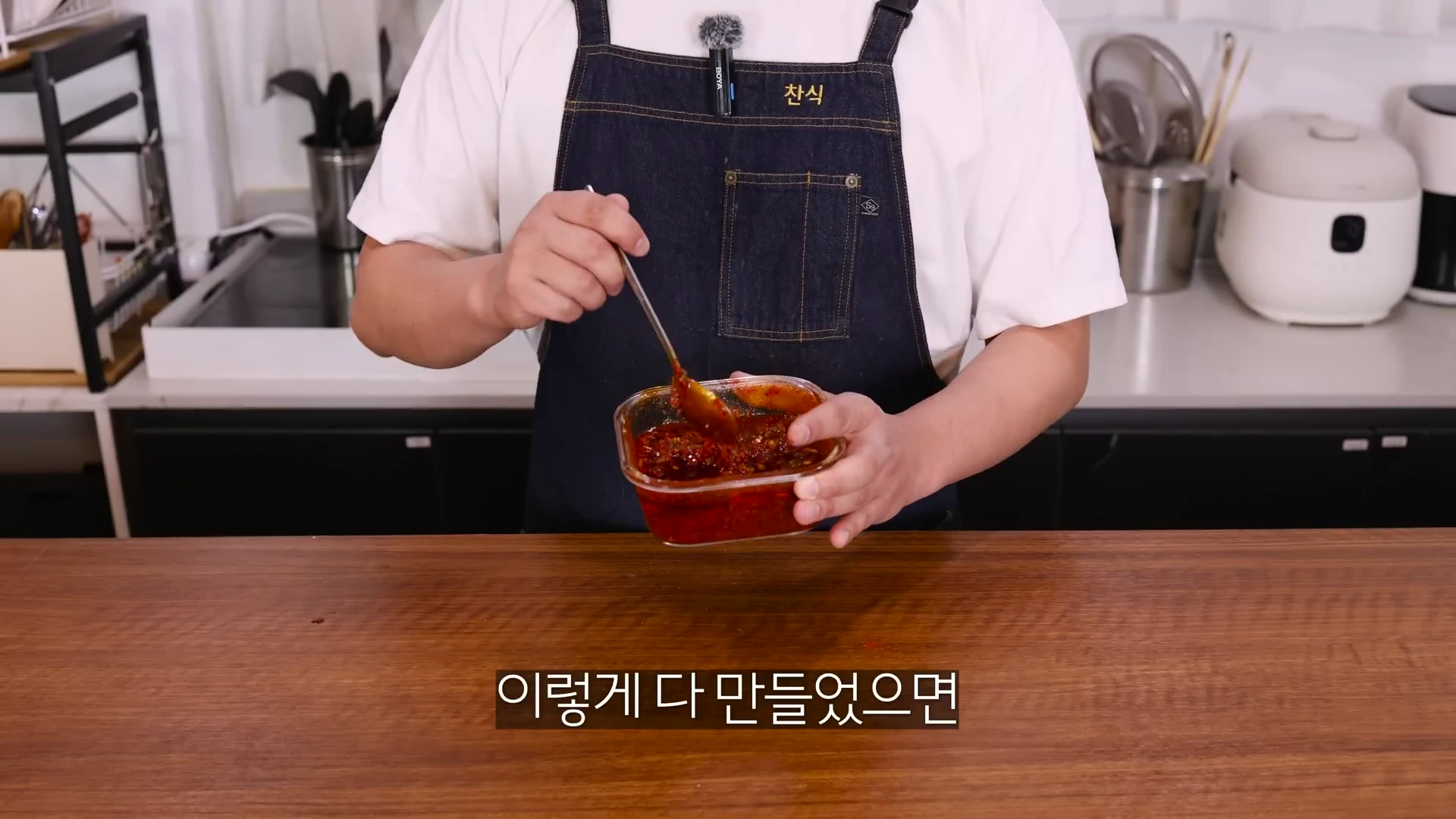 앞치마를 입은 사람이 유리 용기에 담긴 붉은색 고추기름 다대기를 숟가락으로 섞고 있는 모습