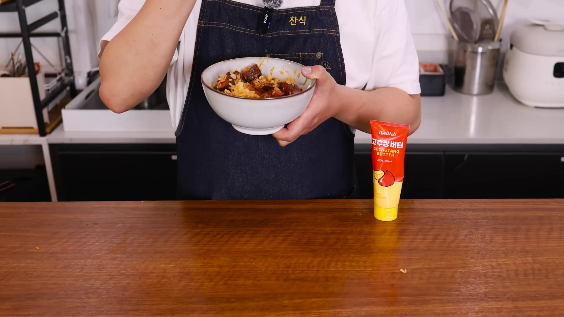 앞치마를 입은 요리사가 스테이크 덮밥이 담긴 그릇을 들고 있고 그 옆에 고추장 버터 튜브가 세워져 있다.