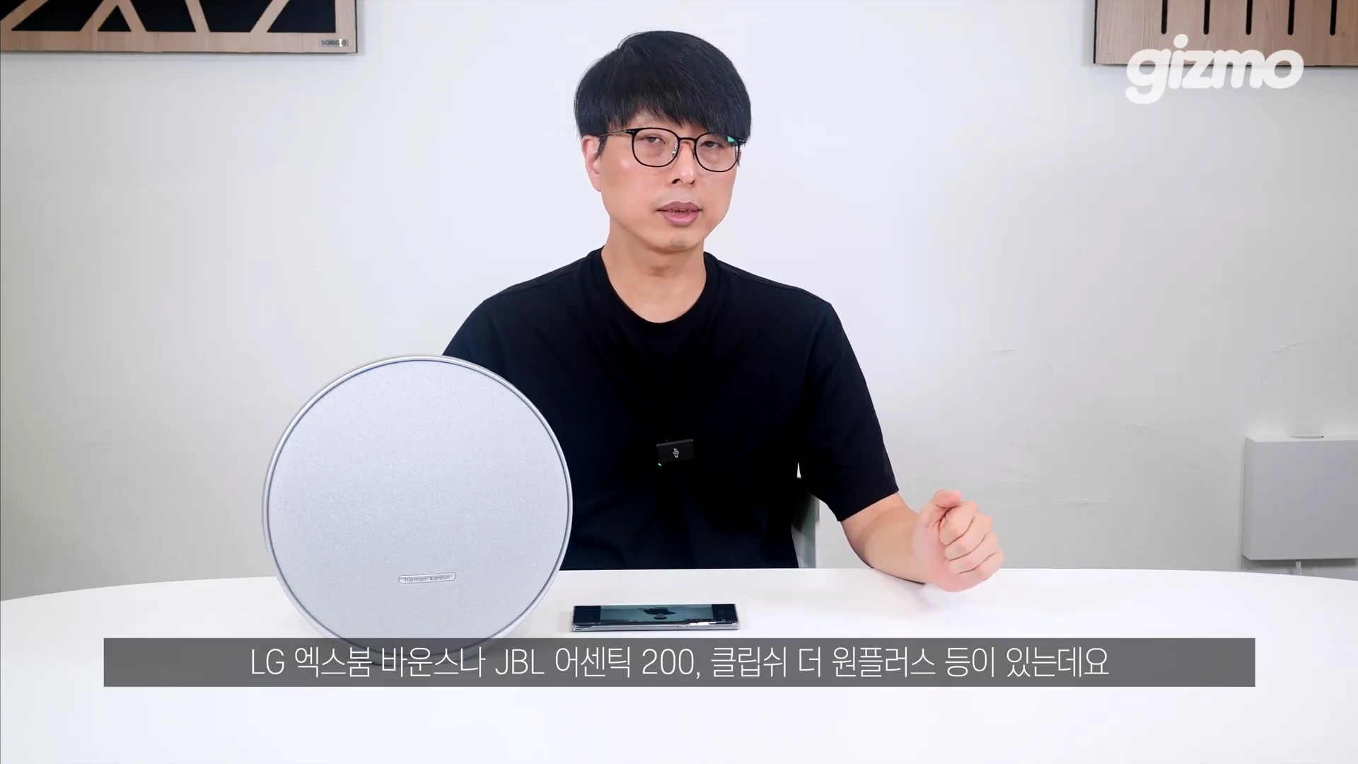 안경을 쓴 남성이 원형 스피커와 스마트폰이 놓인 테이블 앞에 앉아 이야기를 나누고 있는 모습