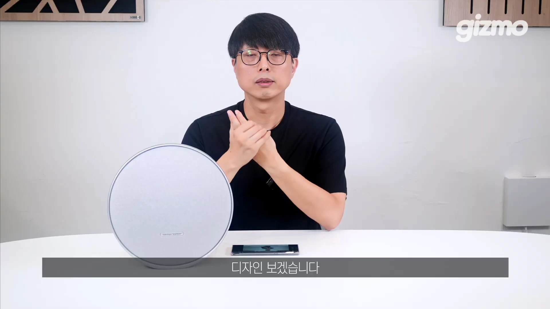 검은색 티셔츠를 입고 안경을 쓴 남성이 스튜디오 테이블 앞에 앉아 원형 스피커를 소개하는 모습