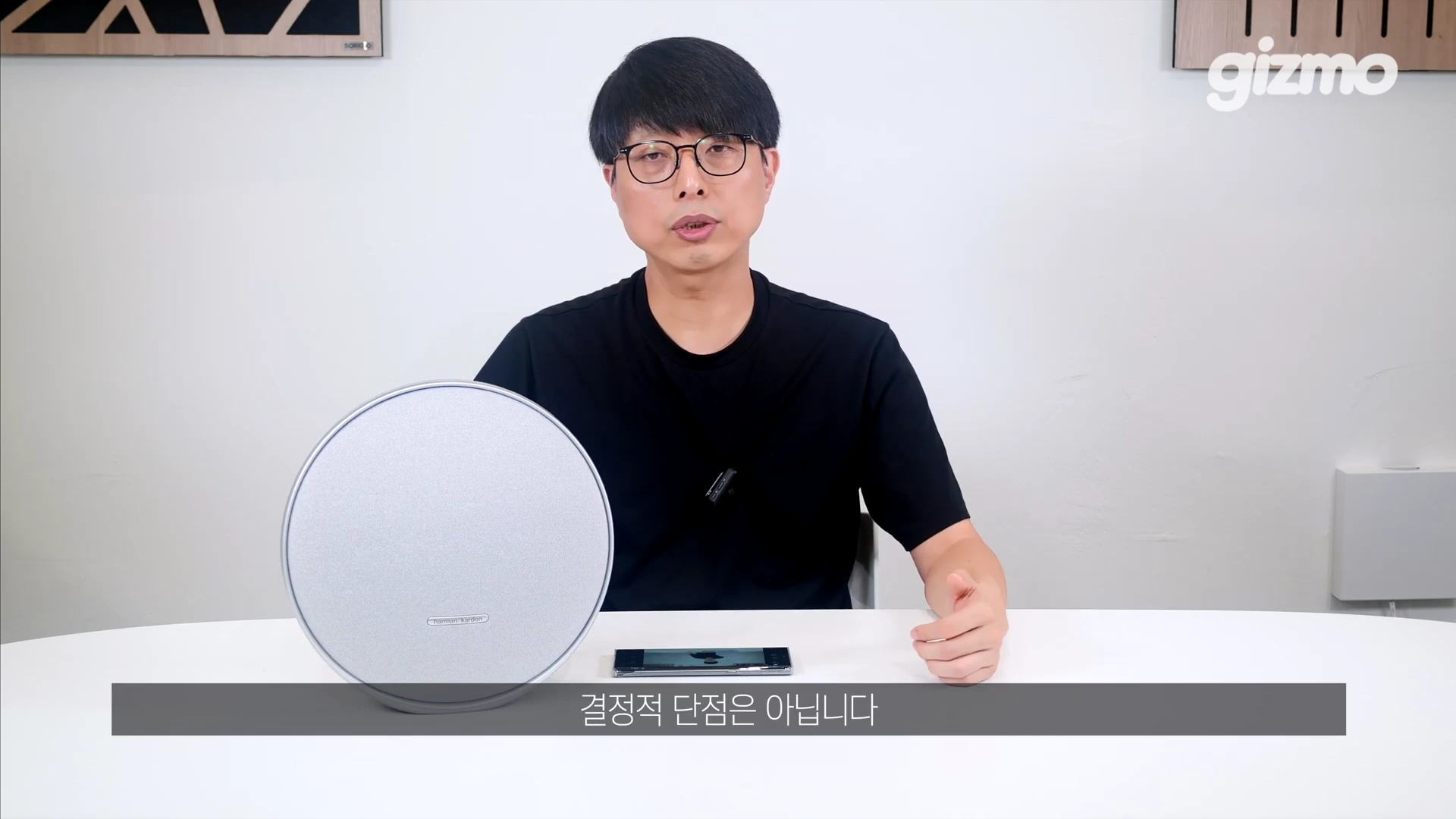 안경을 쓴 남성이 흰색 테이블 위에 놓인 원형 스피커를 앞에 두고 앉아 이야기를 나누고 있는 모습이 담긴 영상 화면
