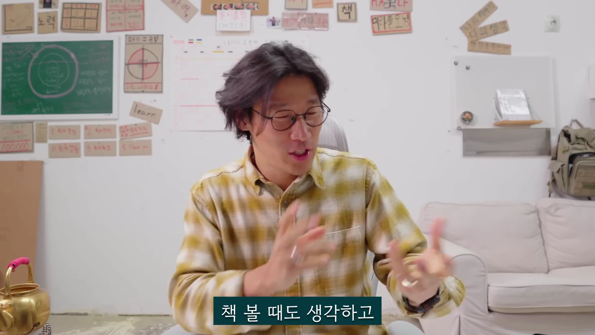 안경을 쓴 남성이 노란색 체크무늬 셔츠를 입고 의자에 앉아 손동작을 하며 이야기를 나누고 있다. 배경 벽면에는 여러 개의 메모와 그림이 붙어 있다.