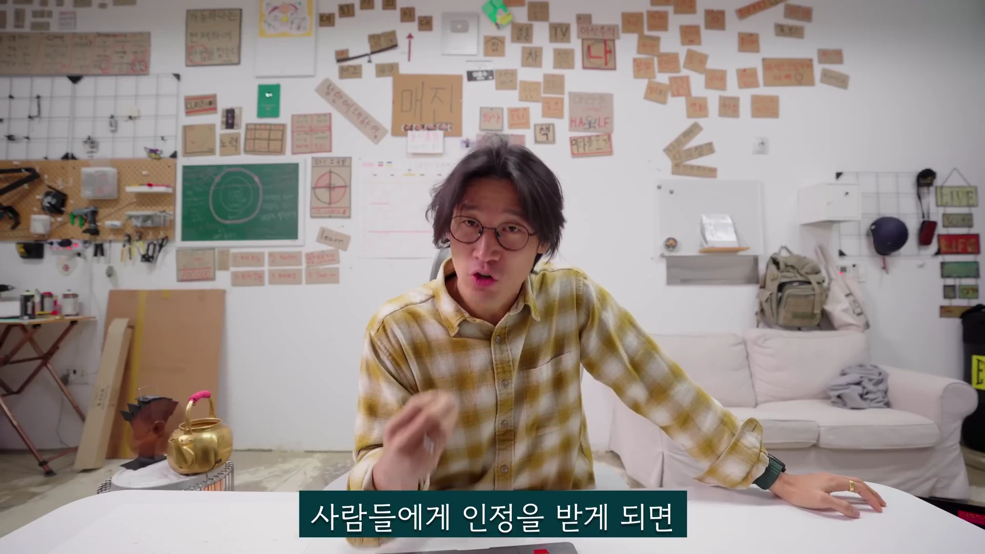 체크무늬 셔츠를 입고 안경을 쓴 남성이 흰색 테이블 뒤에 앉아 손짓을 하며 무언가를 설명하고 있다. 배경의 벽면에는 다양한 메모지가 붙어 있고 칠판과 소파가 배치되어 있다. 하단에는 '사람들에게 인정을 받게 되면'이라는 자막이 표시되어 있다.