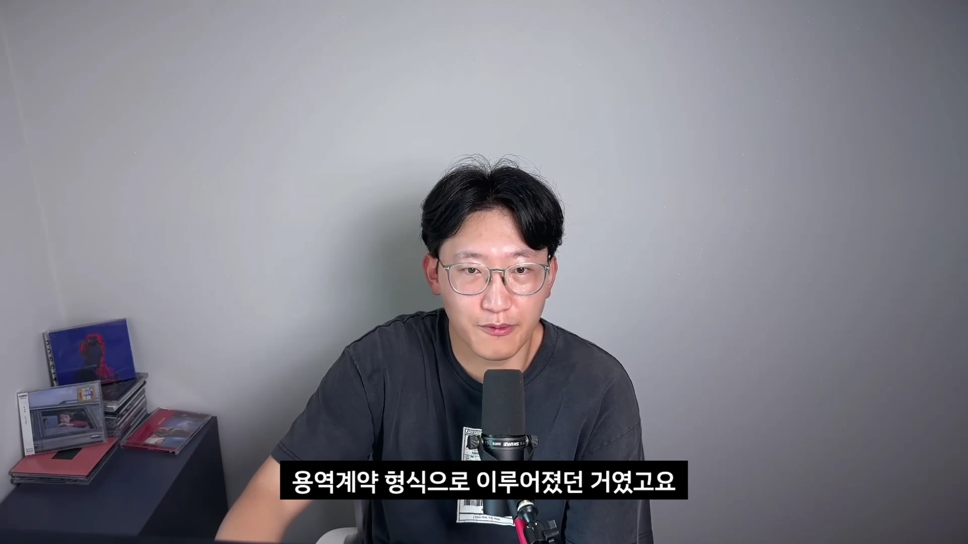 안경을 쓴 남성이 마이크 앞에 앉아 이야기를 하고 있는 모습의 영상 캡처 화면이며, 하단에는 '용역계약 형식으로 이루어졌던 거였고요'라는 자막이 적혀 있다.