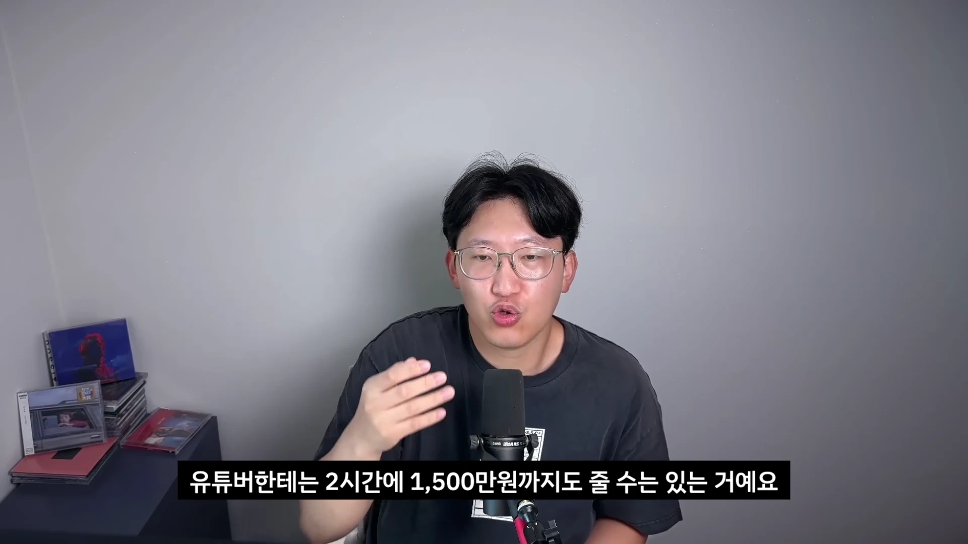 안경을 쓴 한 남성이 회색 배경 앞에서 마이크 앞에 앉아 이야기하는 모습이 담긴 영상 캡처 화면이며, 하단에 '유튜버한테는 2시간에 1,500만 원까지도 줄 수는 있는 거예요'라는 자막이 표시되어 있습니다.