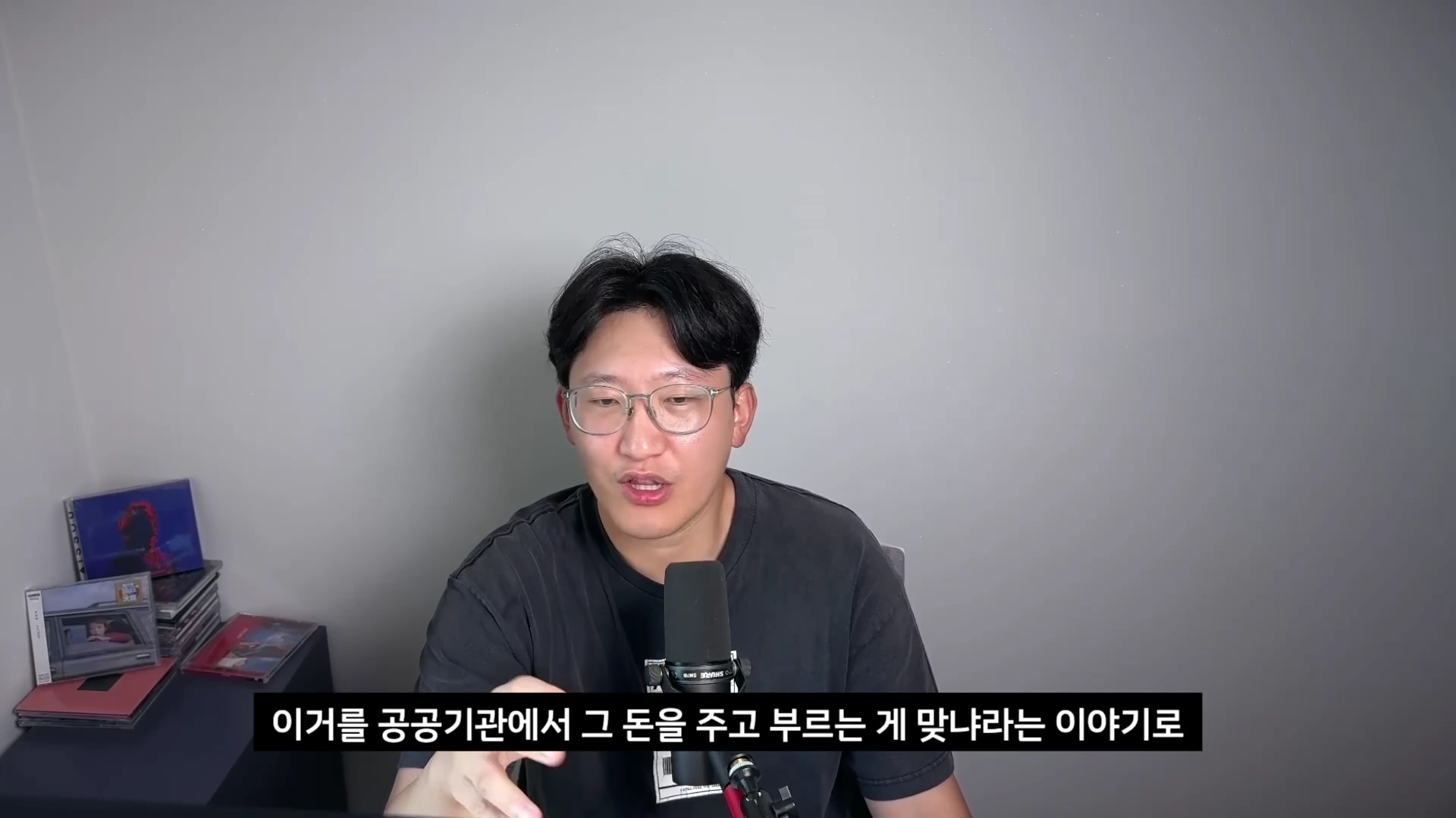 안경을 쓴 남성이 마이크 앞에서 이야기를 하고 있는 모습이 담긴 영상 캡처 화면이며, 화면 하단에는 '이거를 공공기관에서 그 돈을 주고 부르는 게 맞냐라는 이야기로'라는 자막이 표시되어 있다.