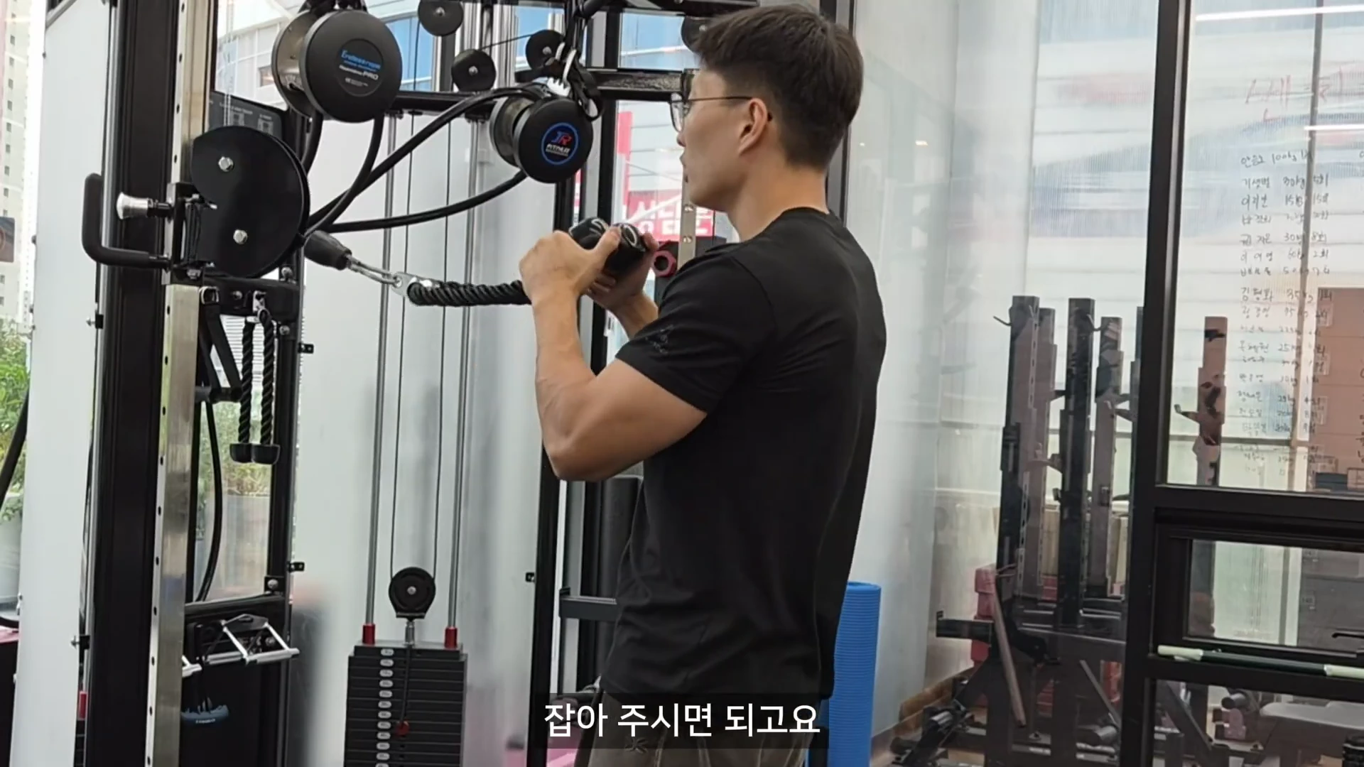 실내 체육관에서 검은색 티셔츠를 입은 남성이 케이블 머신을 사용하여 로프 그립을 당기는 동작을 보여주고 있다.