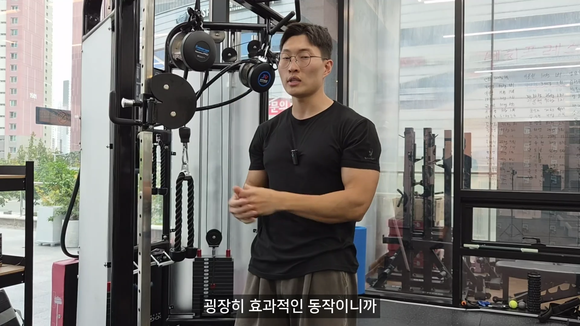 헬스장에서 검은색 반팔 티셔츠를 입고 안경을 쓴 남성이 케이블 머신 앞에 서서 설명하는 모습