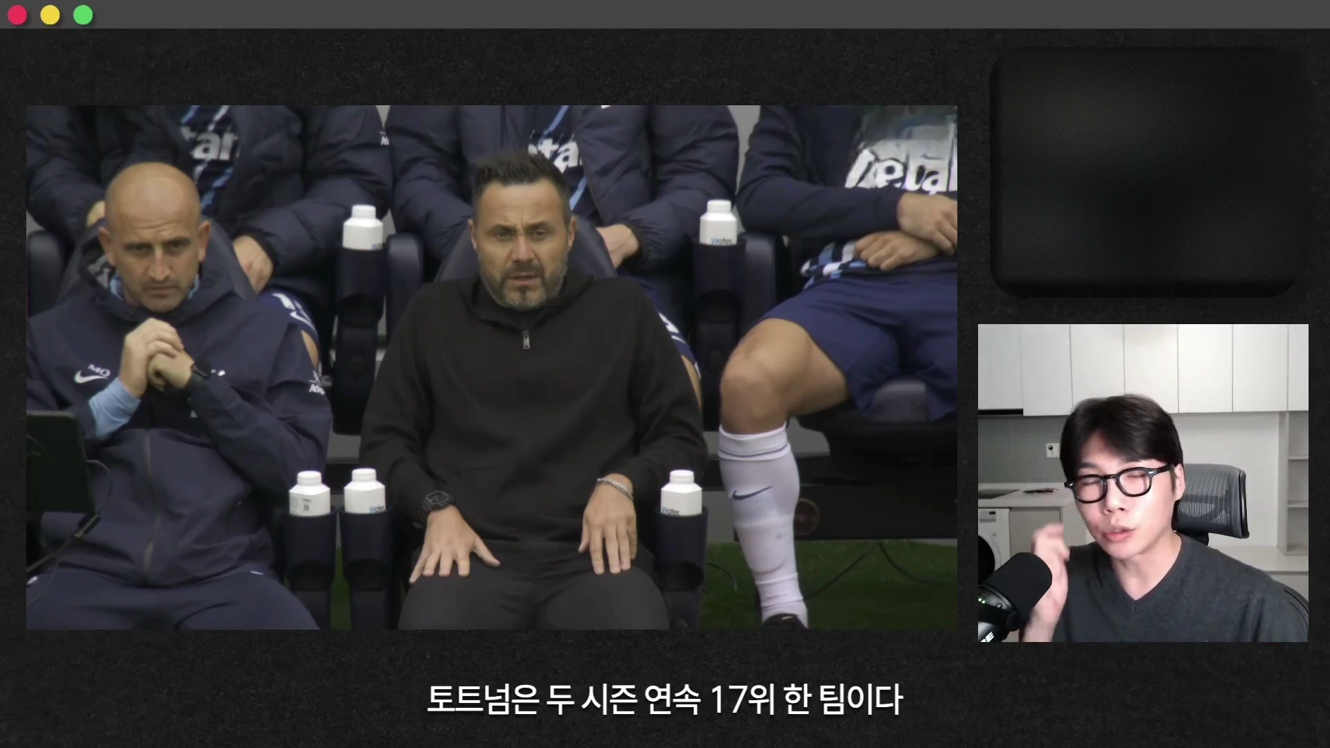축구 경기 중 벤치에 앉아 심각한 표정을 짓고 있는 감독과 코치진의 모습, 그리고 화면 우측 하단에는 안경을 쓴 진행자가 마이크 앞에 앉아 있는 모습이 담긴 화면입니다.