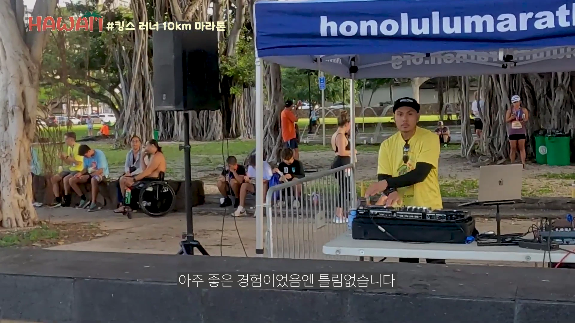 공원 내 야외 행사장 부스에서 노란색 티셔츠를 입은 남성이 디제잉 장비 앞에 서 있는 모습과 주변에 휴식을 취하는 사람들의 풍경