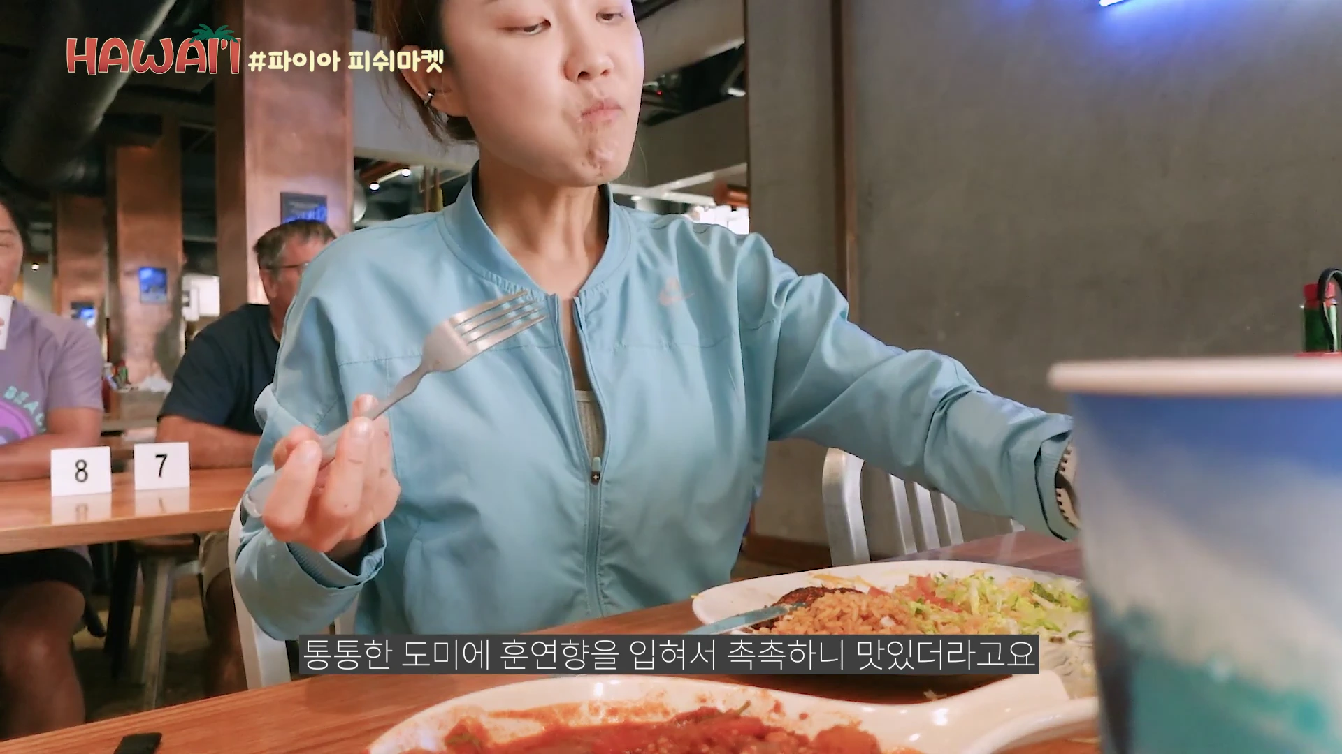 음식점에서 밝은 파란색 바람막이를 입은 여성이 포크를 들고 식사하는 모습