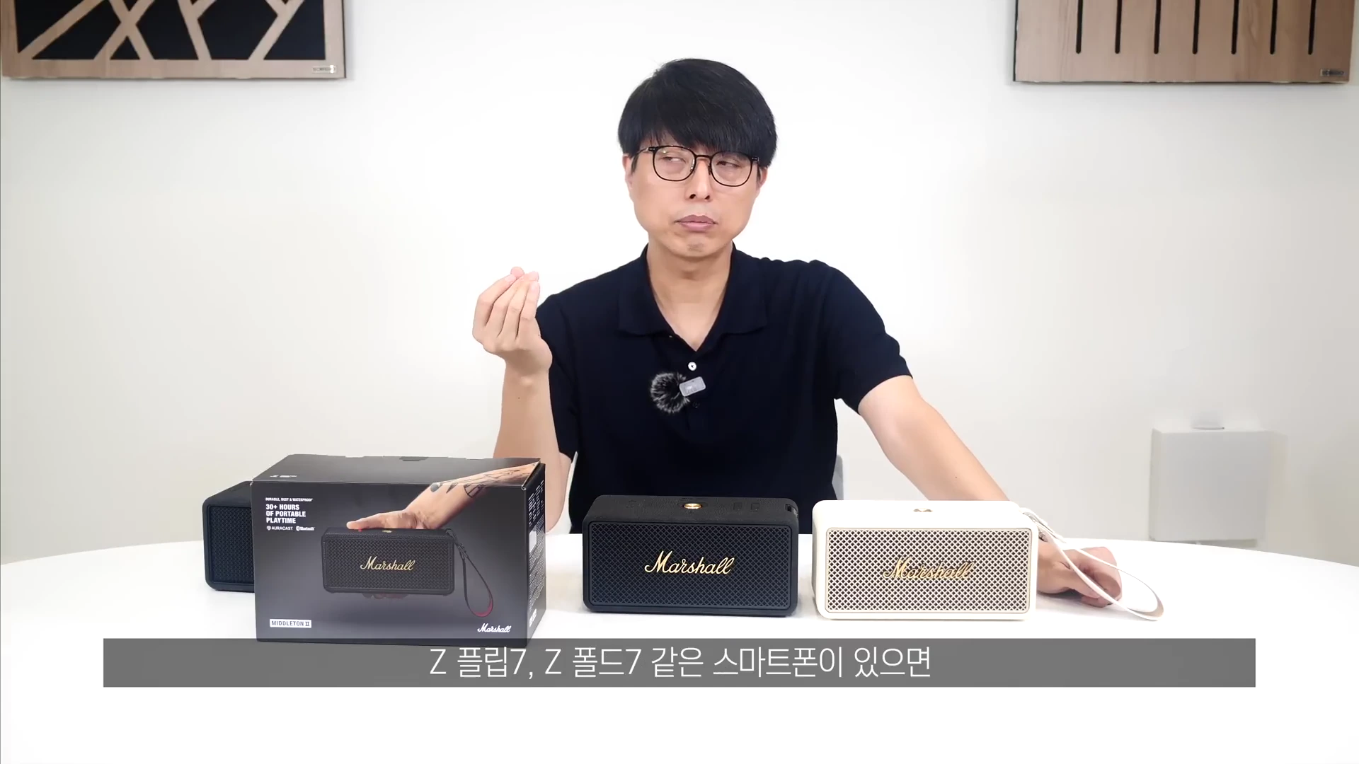 흰색 테이블 위에 마샬 미들톤 2 스피커 블랙과 크림 색상이 제품 박스와 함께 놓여 있고, 안경을 쓴 남성이 손짓하며 설명하고 있는 유튜브 영상의 한 장면입니다.