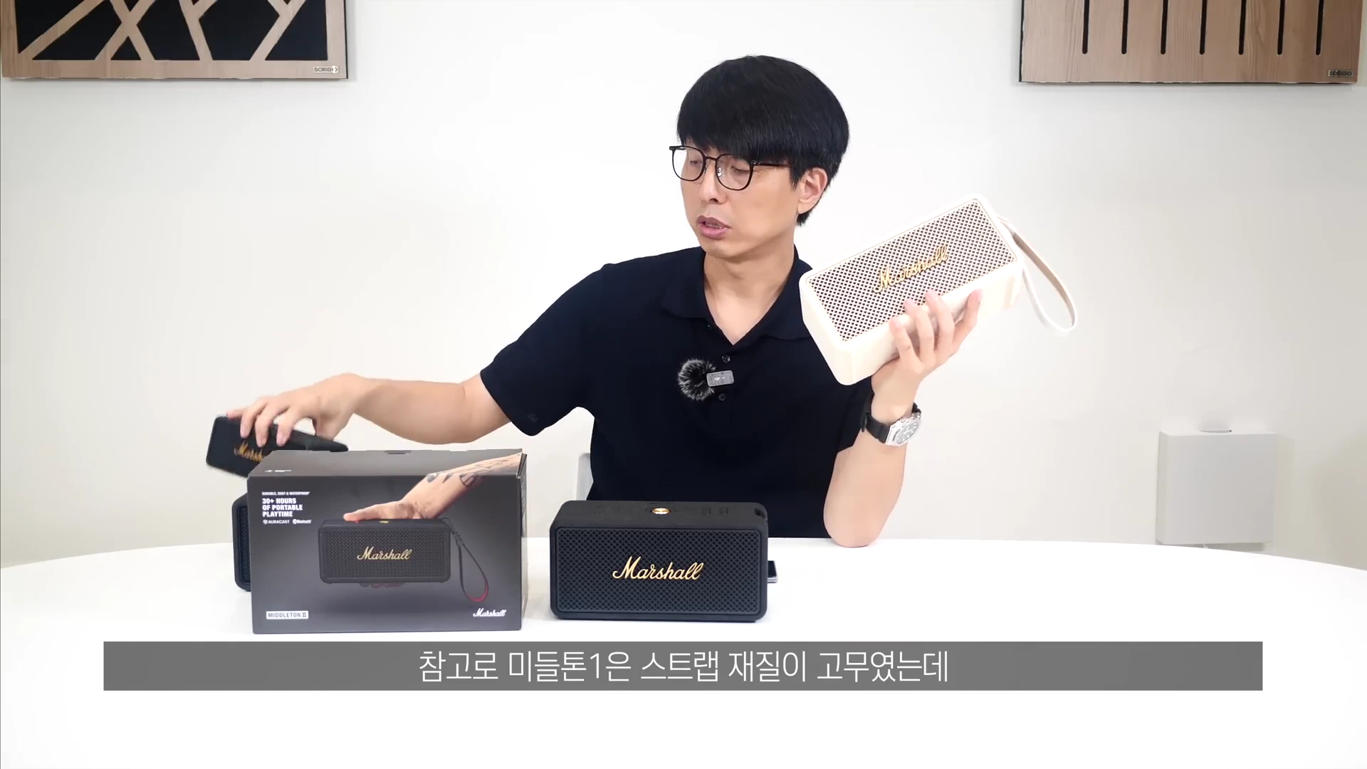 안경을 쓴 남성이 밝은 스튜디오 탁자 위에 놓인 마샬 포터블 스피커를 한 손으로 들어 보이며 다른 손으로는 제품 패키지를 만지고 있는 모습