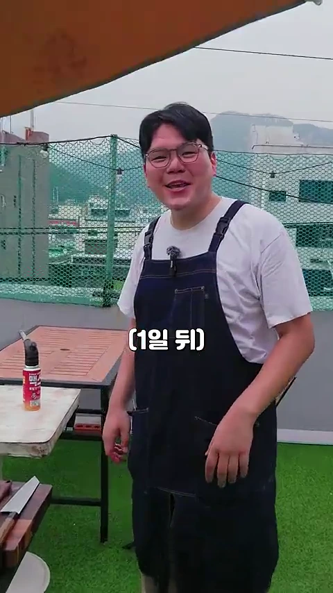 안경을 쓰고 앞치마를 두른 남성이 옥상 야외 공간에서 카메라를 향해 웃으며 서 있고, 화면 중앙에는 '(1일 뒤)'라는 자막이 표시되어 있다.