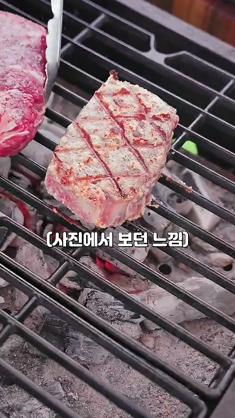 그릴 위에 놓인 스테이크 표면에 다이아몬드 격자무늬의 구운 자국이 선명하게 새겨져 있고 집게가 고기 옆에 위치해 있다.